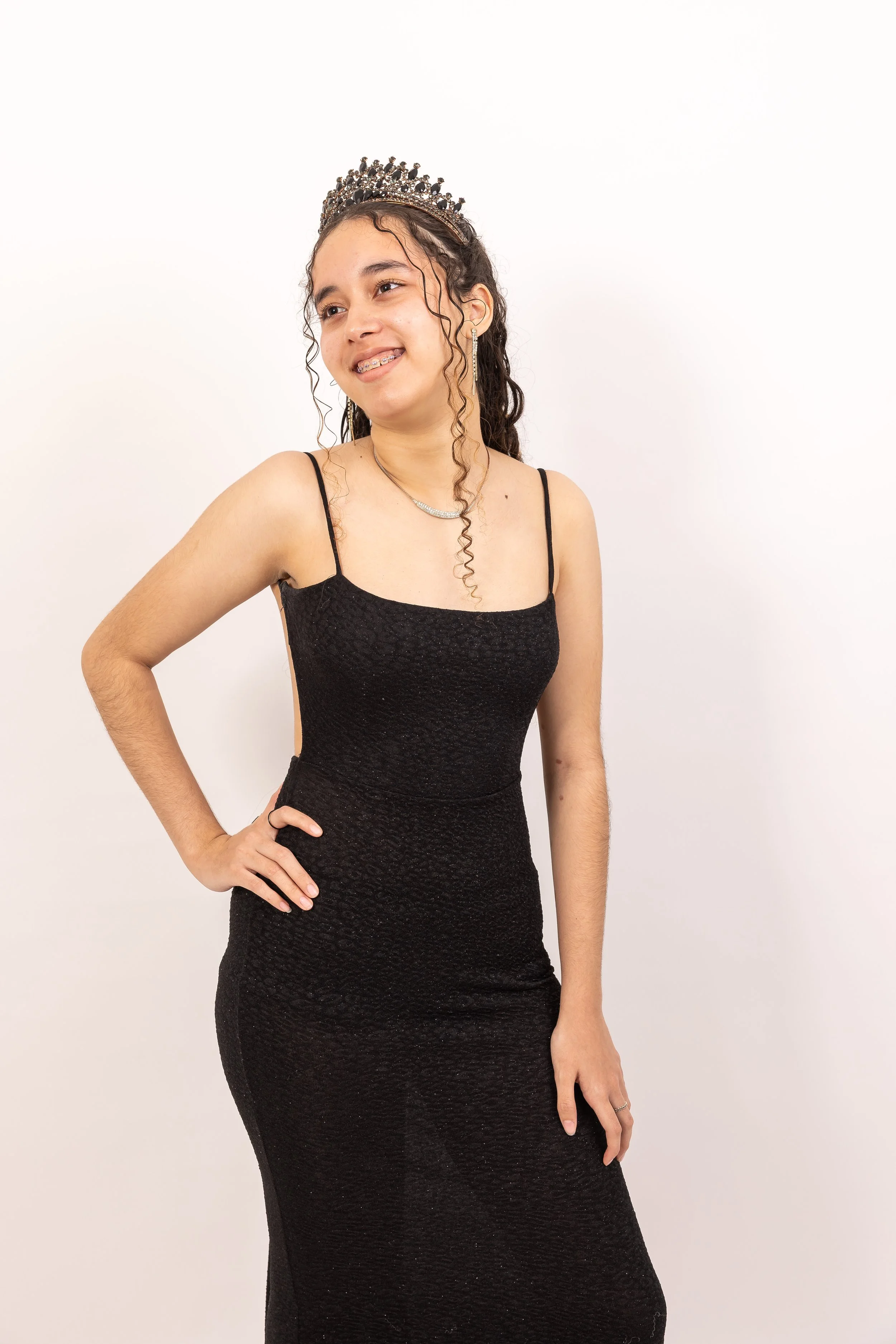 Juvenil mujer con vestido negro ajustado, tiara y joyas, sonriendo y posando con una mano en la cadera, en fondo blanco.