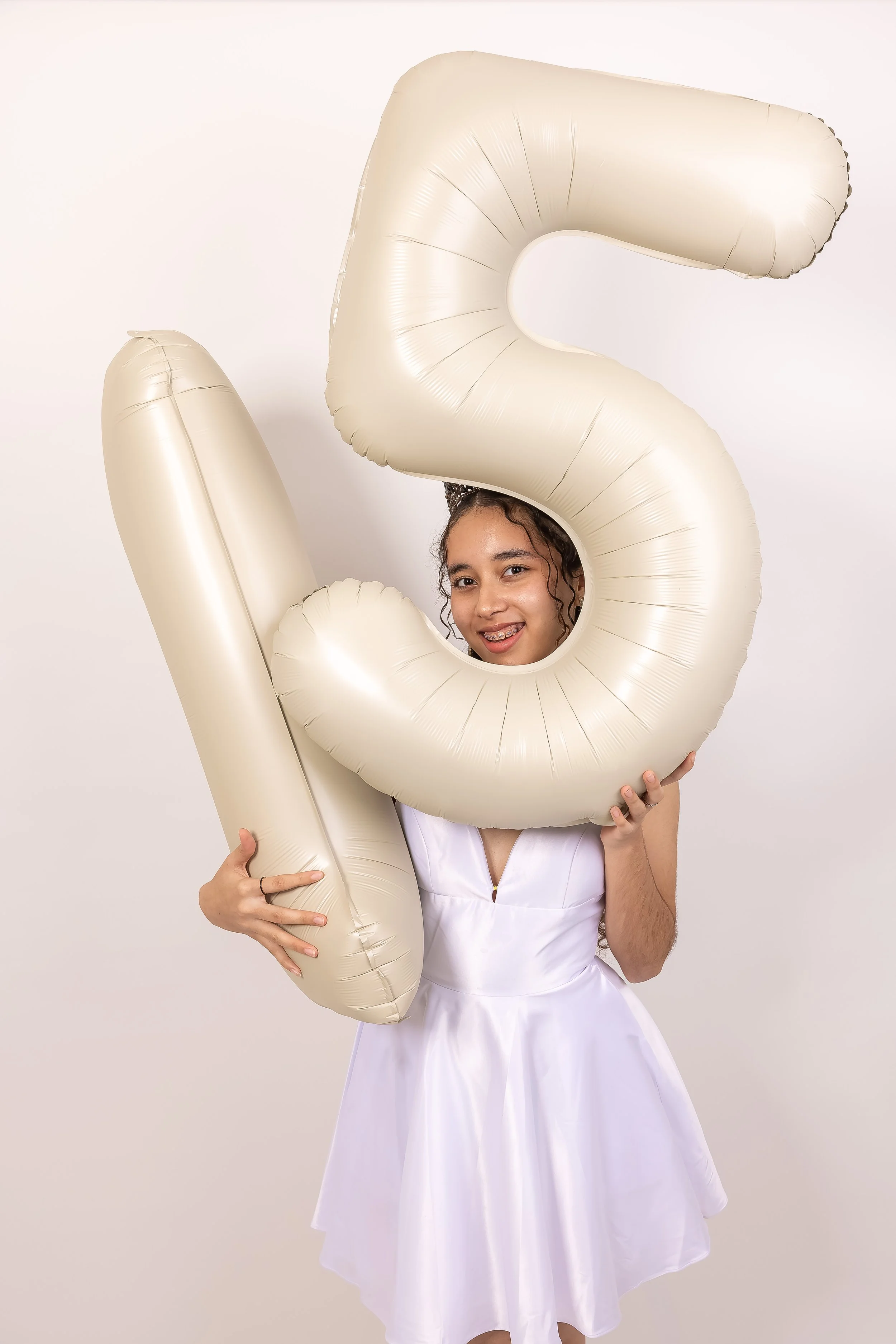 Joven con vestido blanco sosteniendo globos en forma de número 5, en fondo blanco.