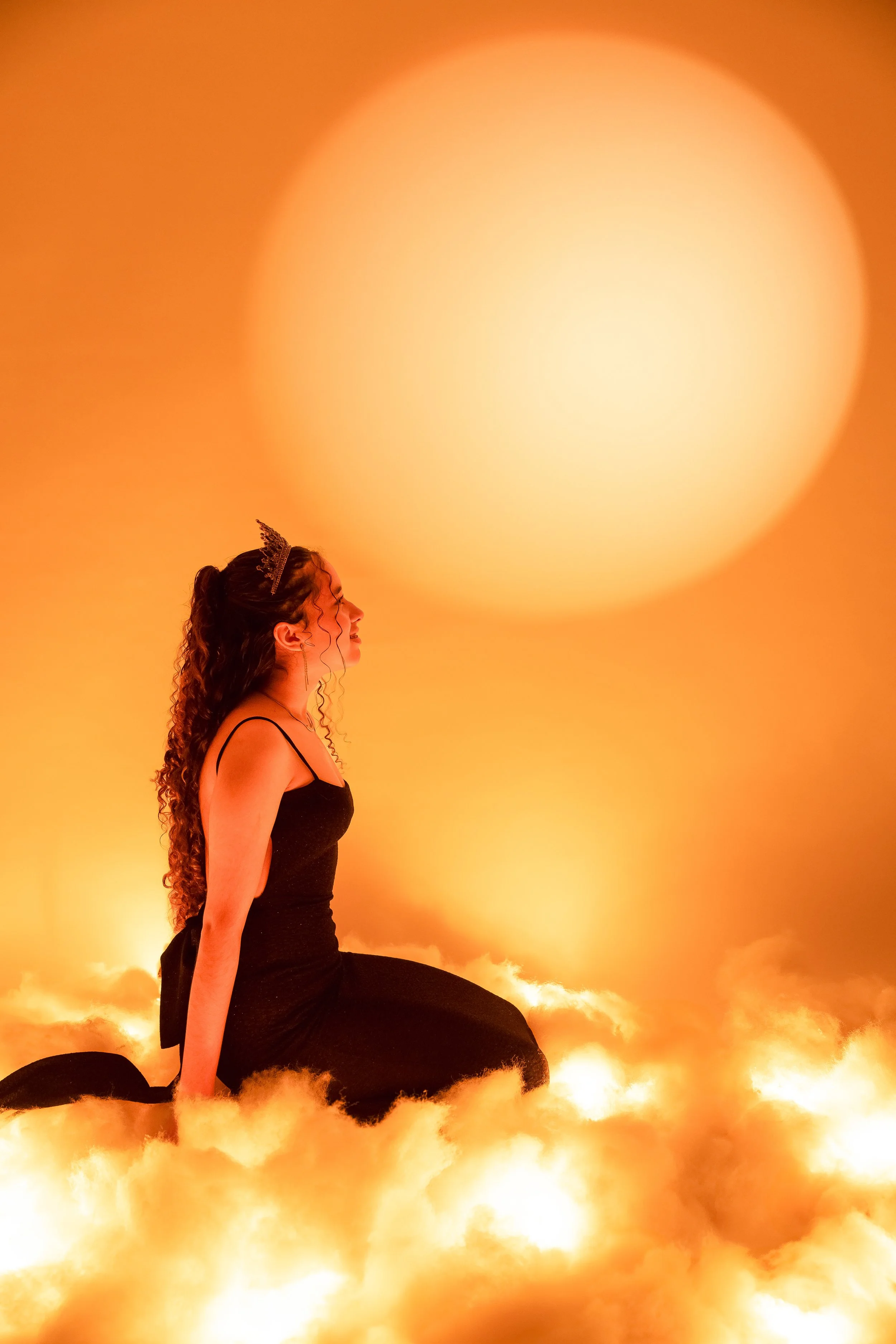 Mujer con cabello rizado y diadema, vestida de negro, arrodillada en nubes de color naranja, con una gran luz amarilla en el fondo.