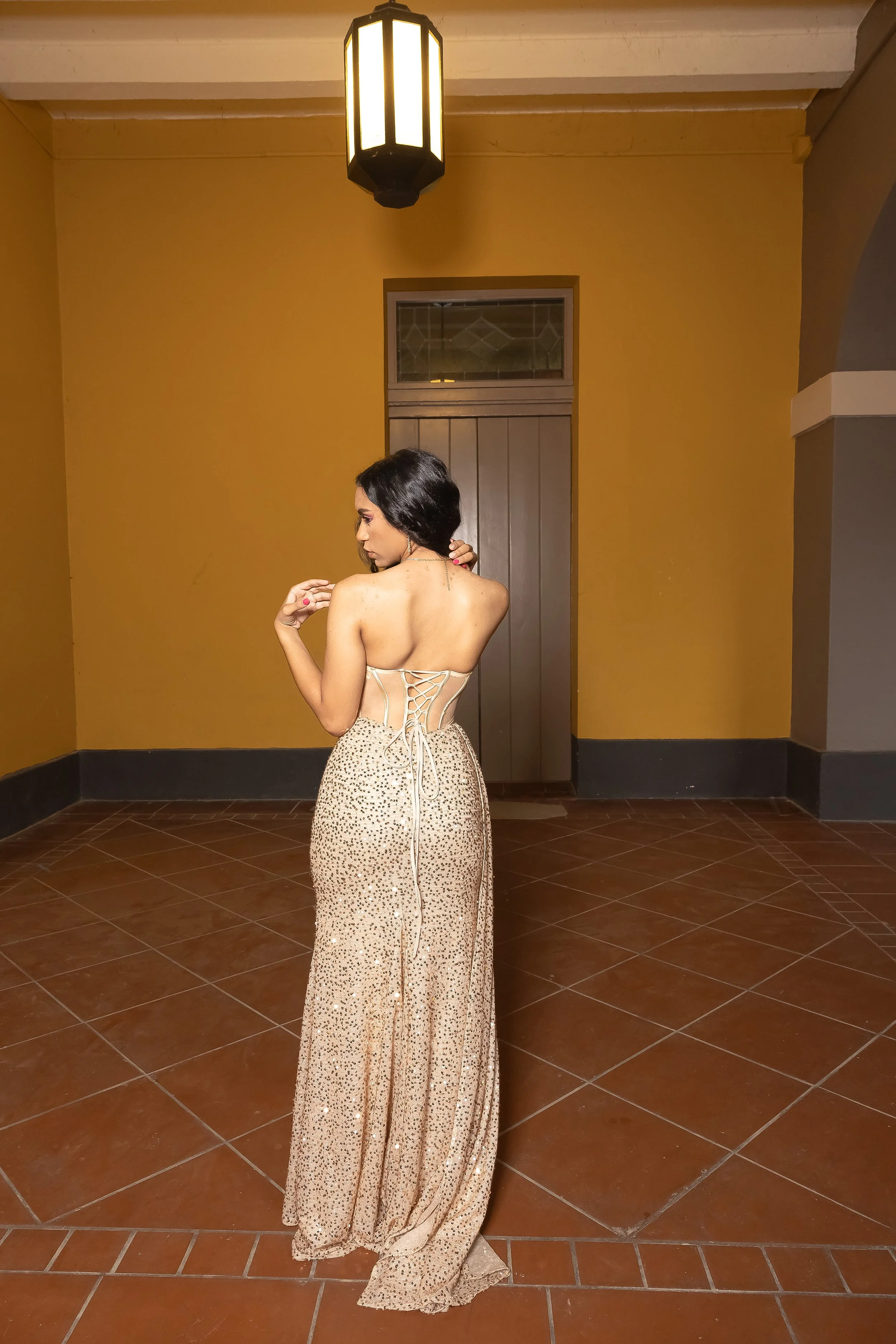 Mujer vestida con vestido largo y brillante, de espaldas en un espacio con paredes naranjas y piso de cerámica, iluminación cálida.