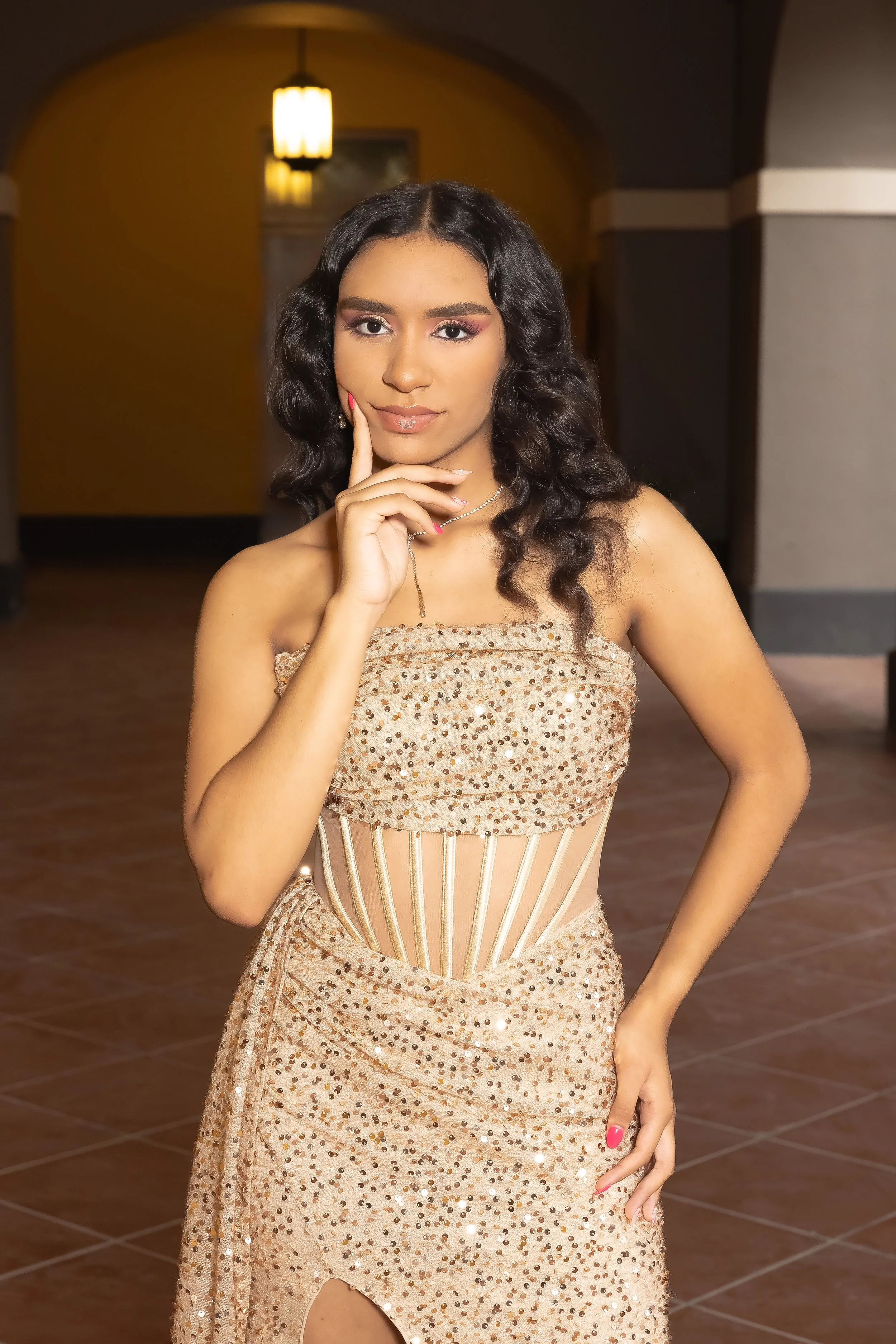 Joven mujer con vestido dorado con detalles de lentejuelas y estructura en el centro, posando en un espacio con iluminación cálida y paredes con colores oscuros y claros.