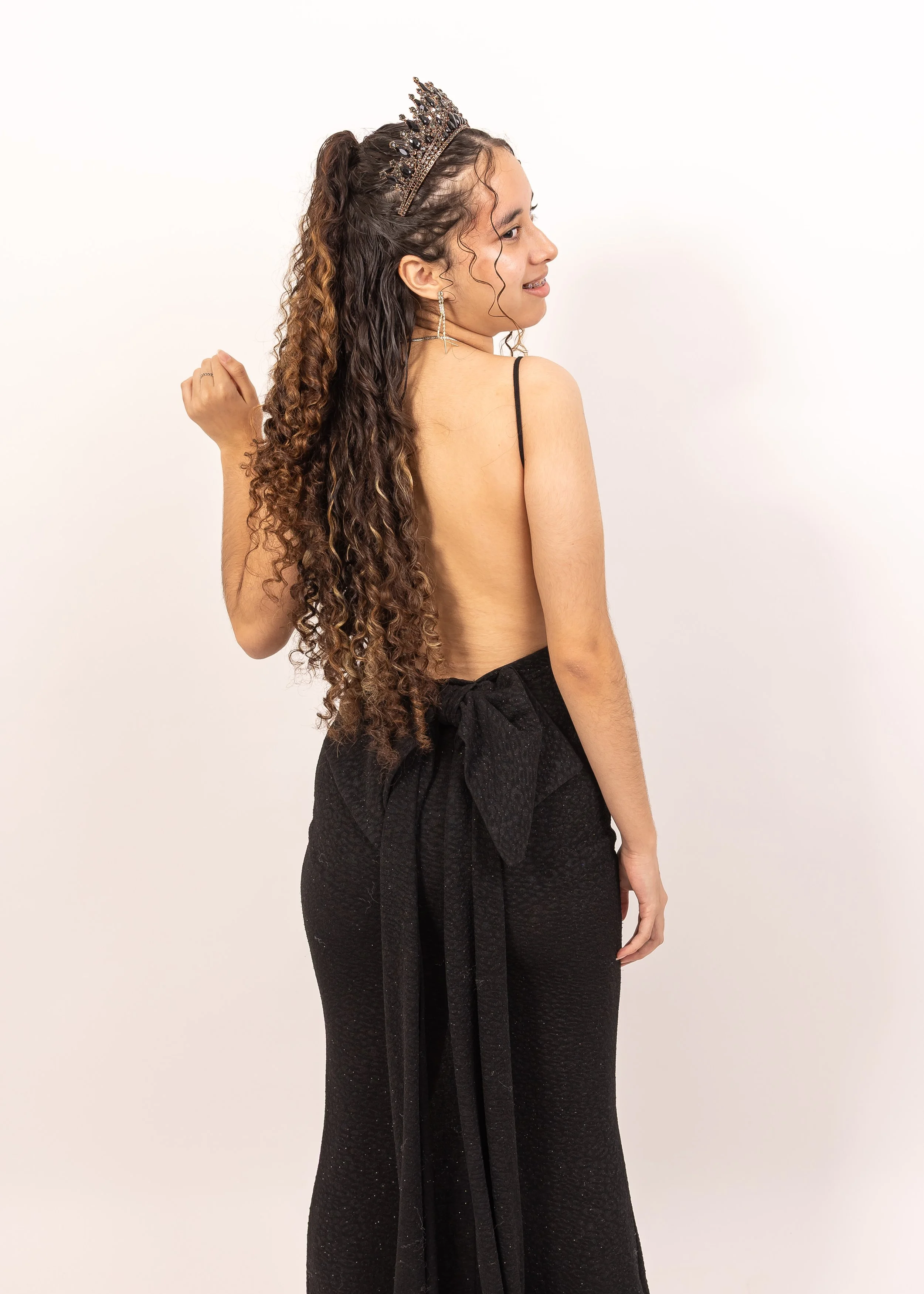 Una mujer con cabello rizado, llevando una tiara y pendientes grandes, viste un vestido negro abierto en la espalda y con lazos en la cintura, posando de lado frente a un fondo blanco.