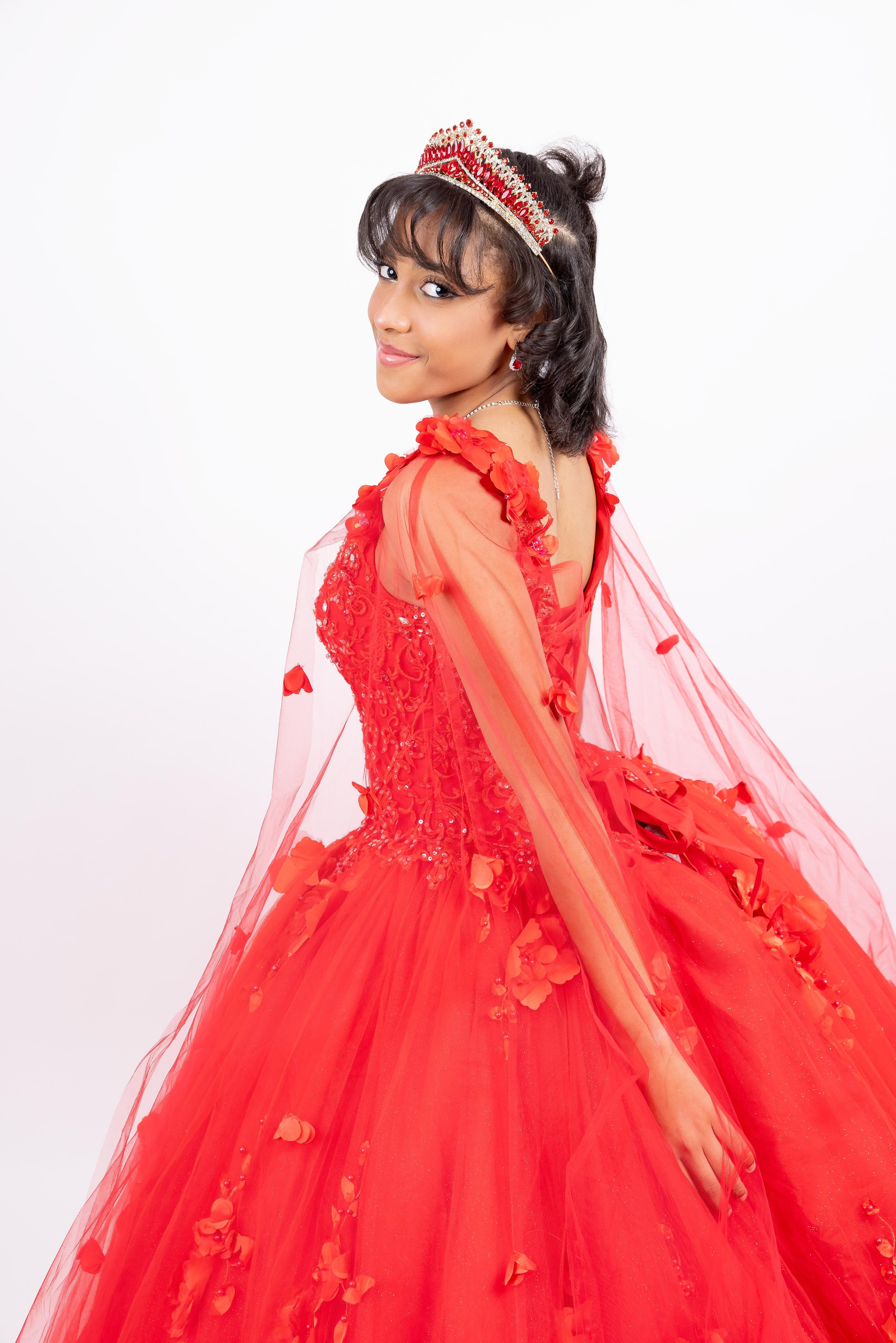 Joven con vestido de fiesta rojo, corona y accesorios, posando de lado en estudio con fondo blanco.
