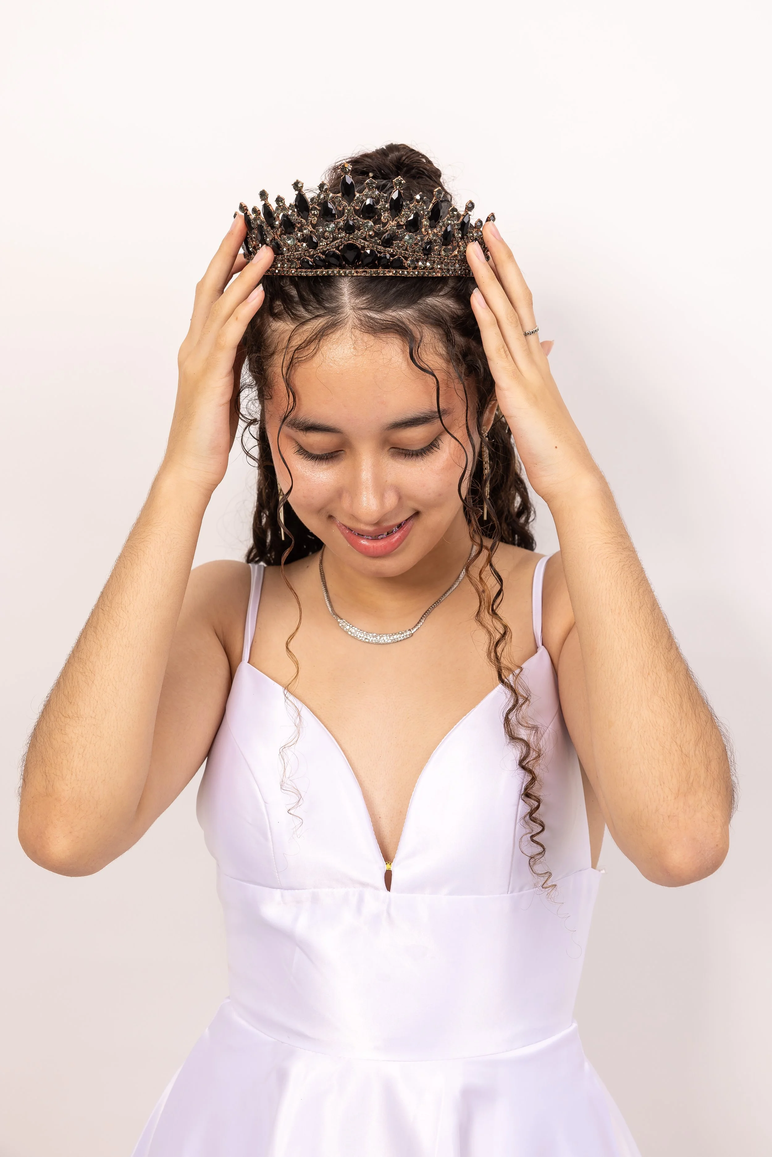 Joven mujer con vestido blanco y corona de princesa, sonriendo y mirando hacia abajo en un fondo blanco.