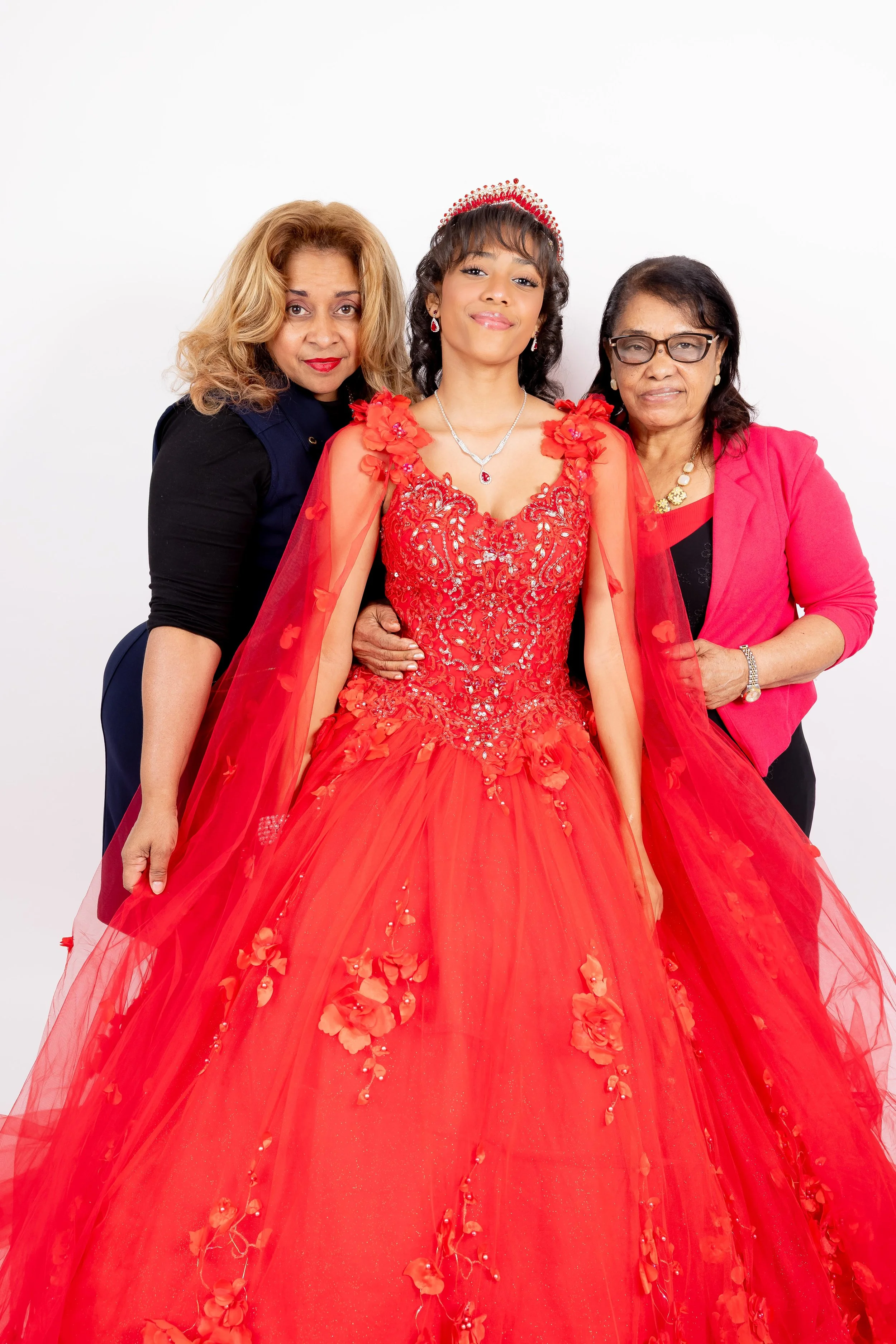 Tres mujeres posando, una en el centro vestida con un vestido largo y rojo con detalles brillantes, y las otras dos a los lados, una con blusa negra y chaqueta rosa, y la otra con blusa negra y chaqueta fucsia, en un fondo blanco.
