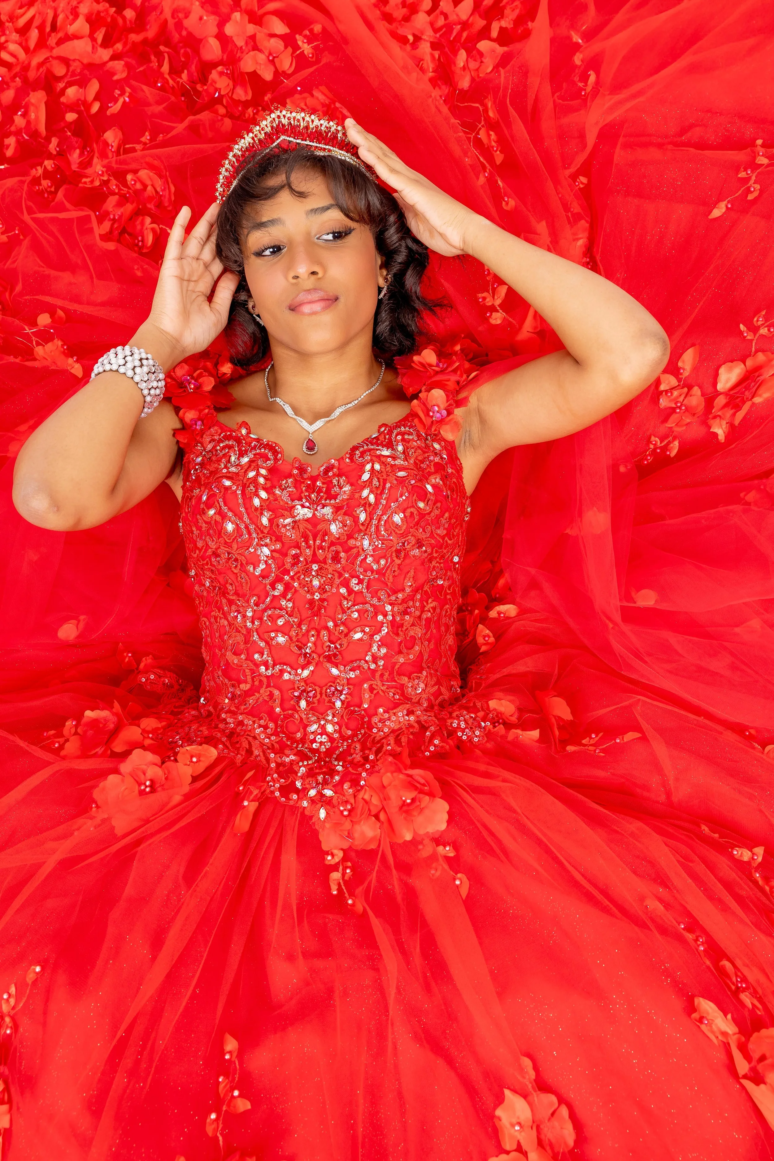 Mujer con vestido rojo de noche, diadema, collar y pulsera, recostada sobre una cama de pétalos de flores rojas.