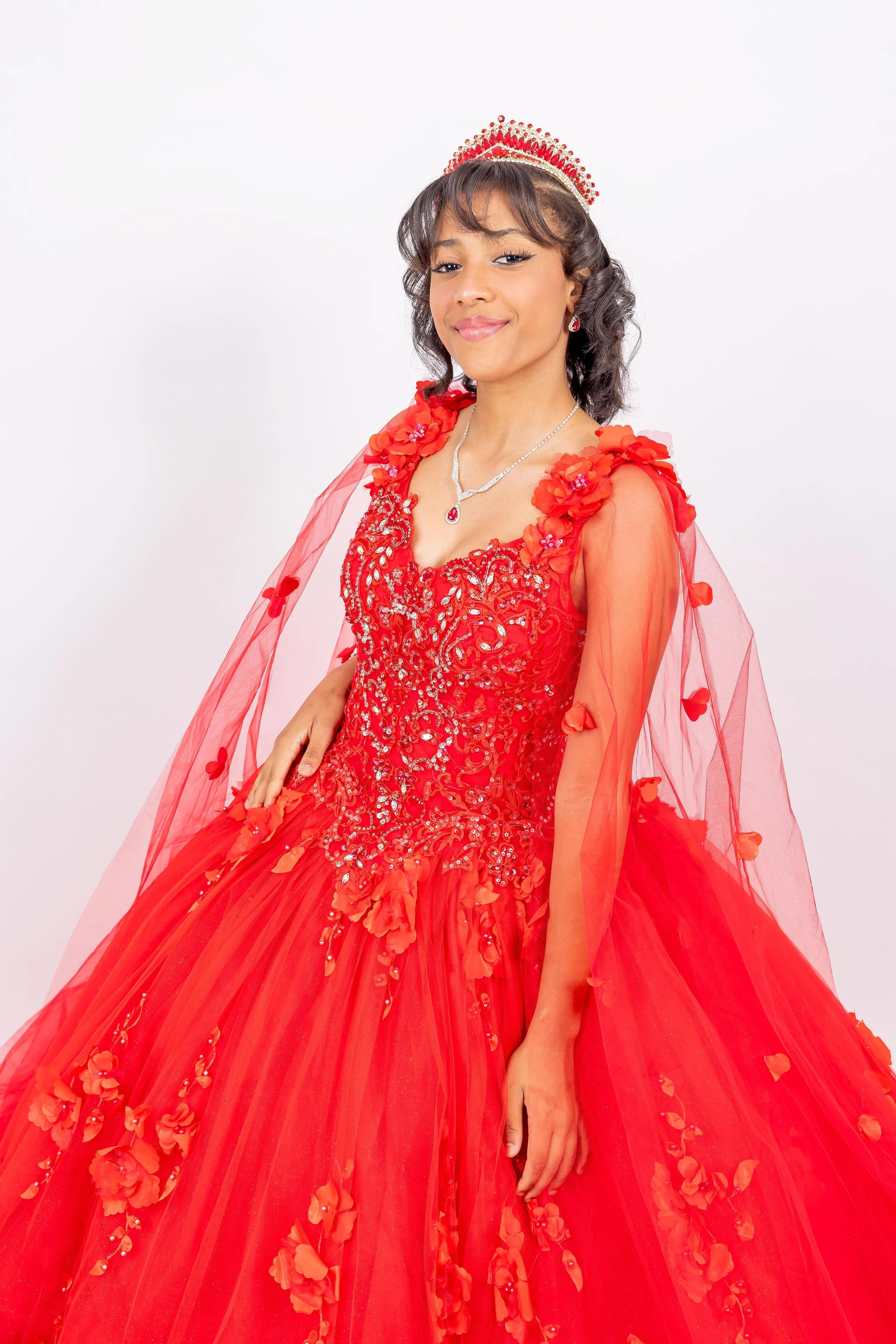 Juvenil con vestido de fiesta rojo con detalles de flores y cristales, usando una tiara, collar y pendiente, sonriendo en un fondo blanco.