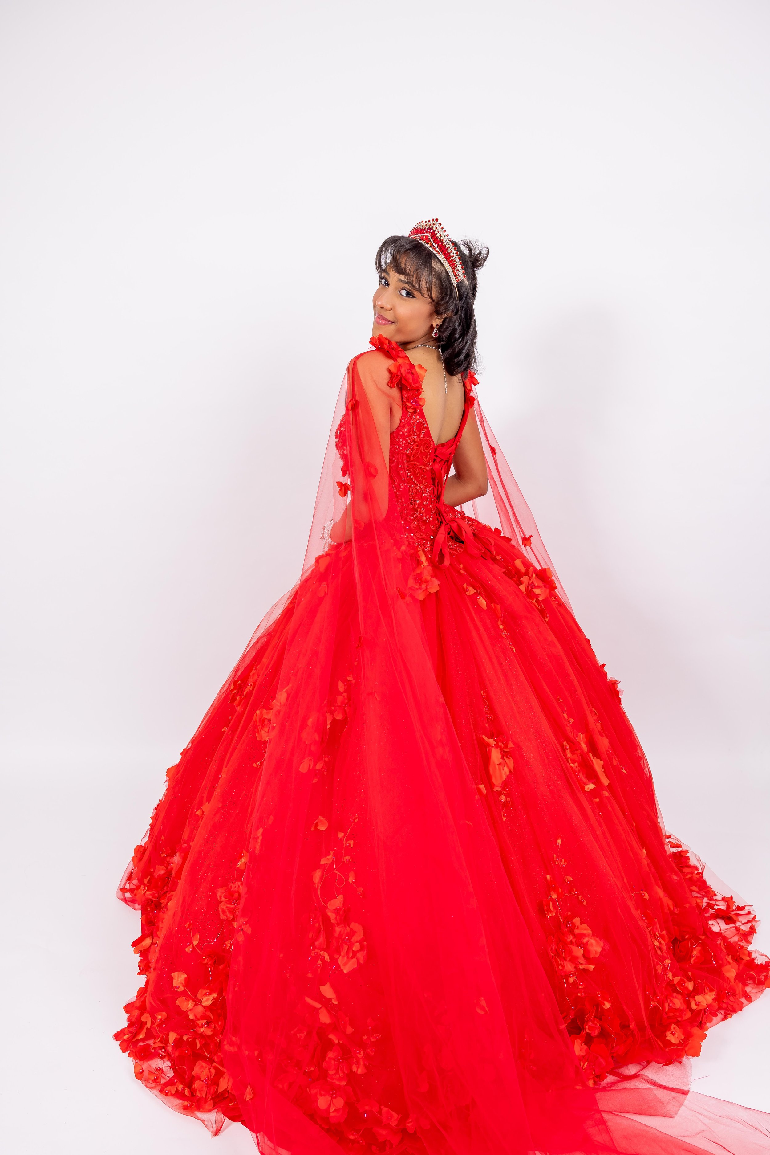 Joven vestida con un vestido largo y rojo, con detalles en tul y flores, usa una tiara y está de espaldas, sonriendo ligeramente.
