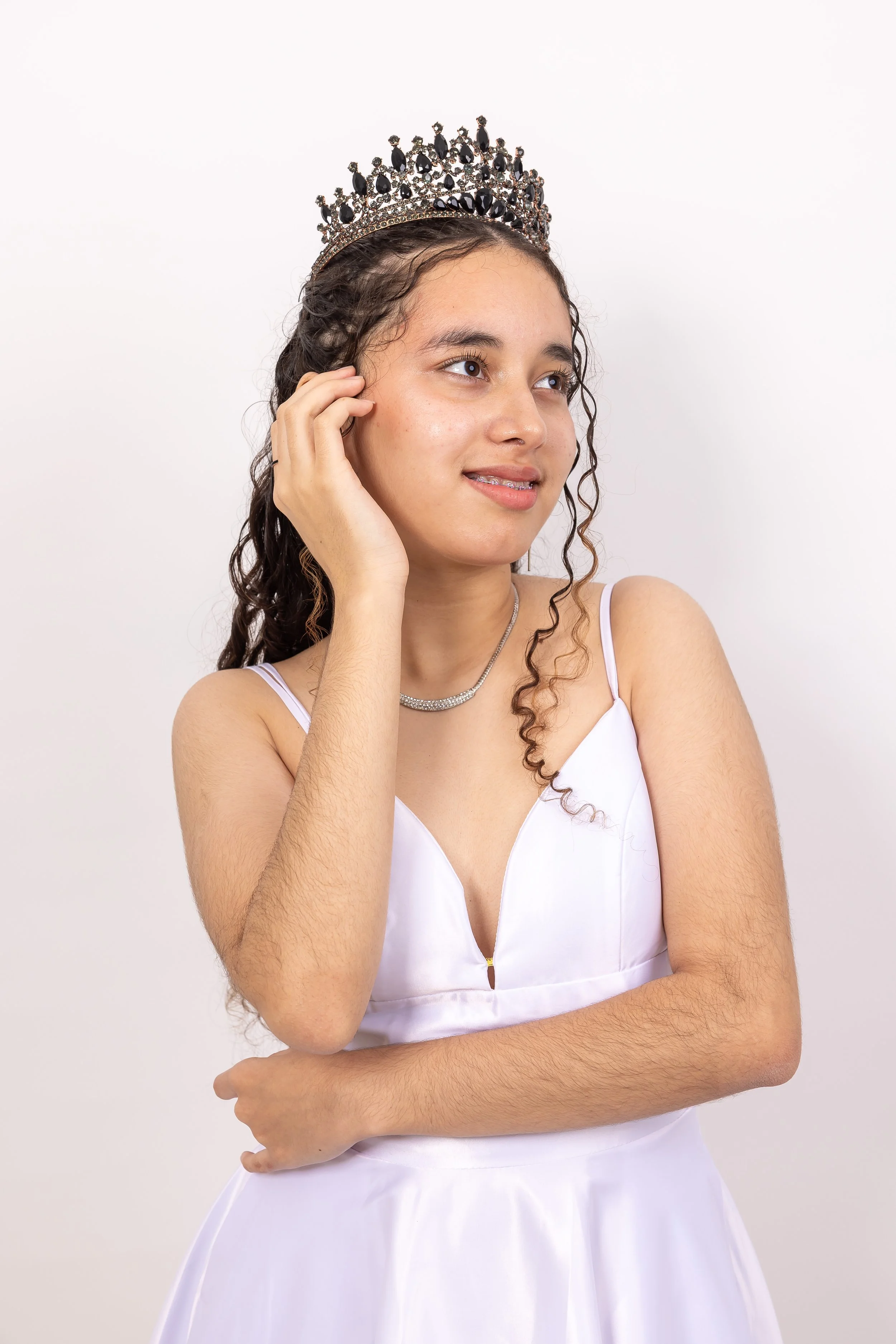 Niña joven con corona y vestido de gala blanco, posando con una mano en la cara y la otra cruzada, sonrisa suave, fondo blanco.