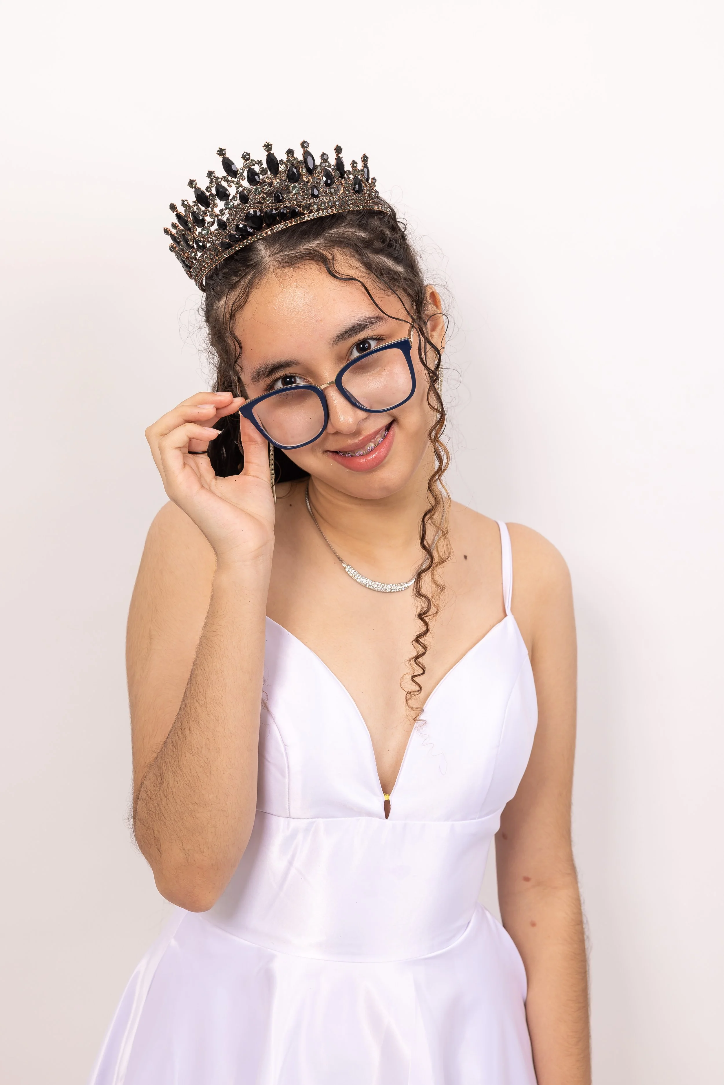 Joven con vestido blanco, diadema de corona y gafas grandes, sonriendo y ajustando sus gafas, contra fondo blanco.