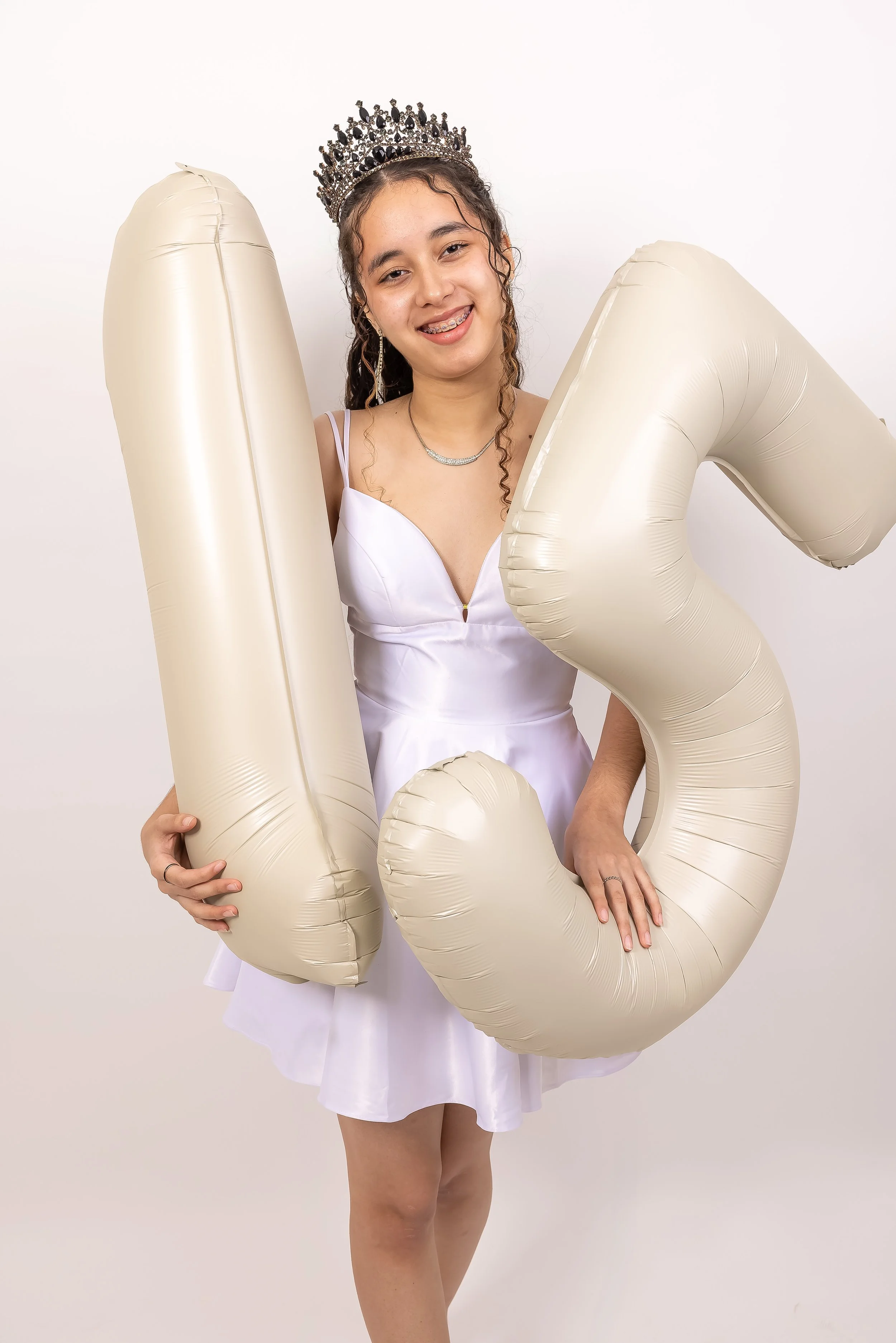Joven con corona y vestido blanco, sosteniendo globos en forma de la letra 'L' y el número '3', sonriendo y de fondo blanco.