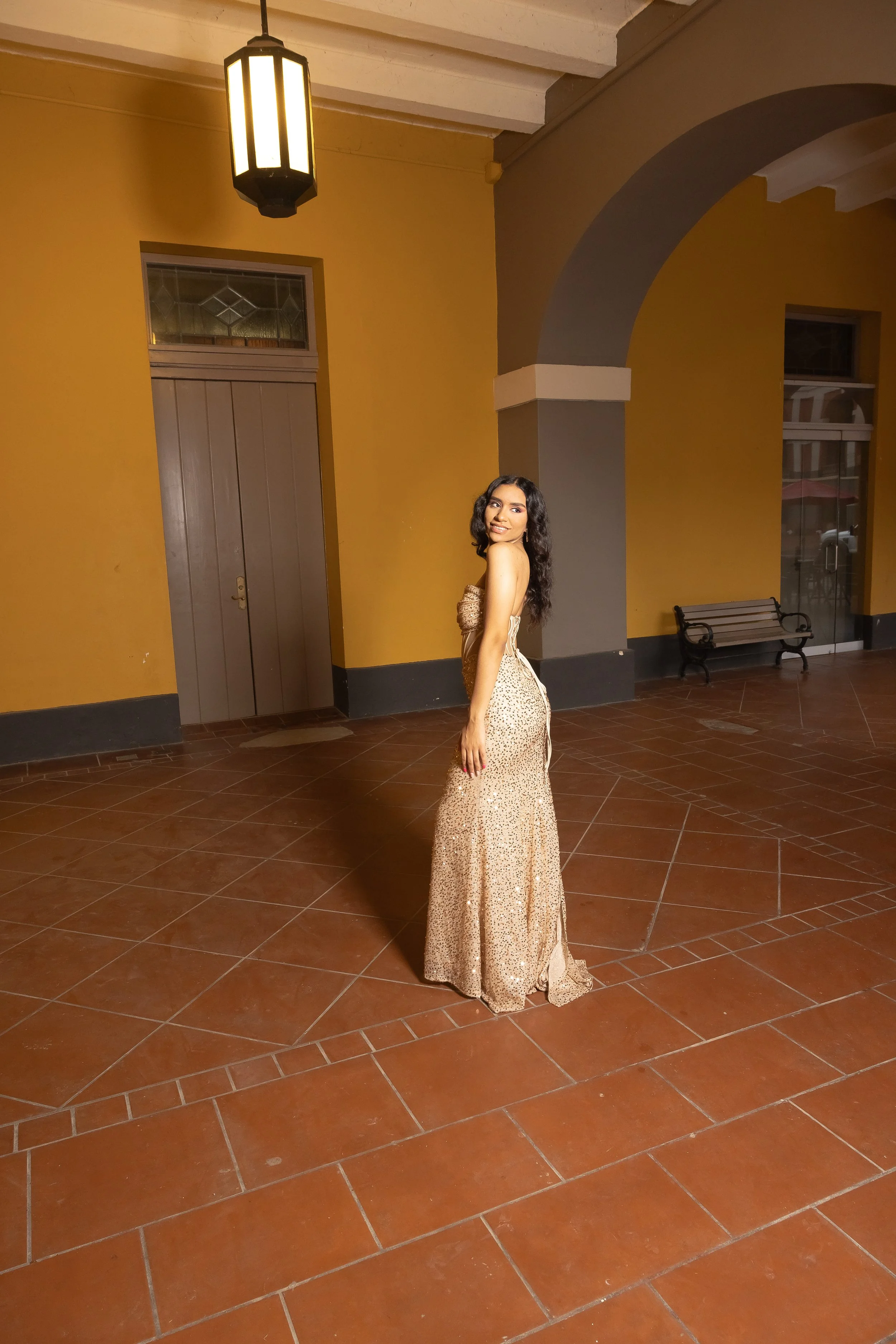Joven mujer con vestido largo de lentejuelas doradas en un espacio con paredes naranjas y ventanas.