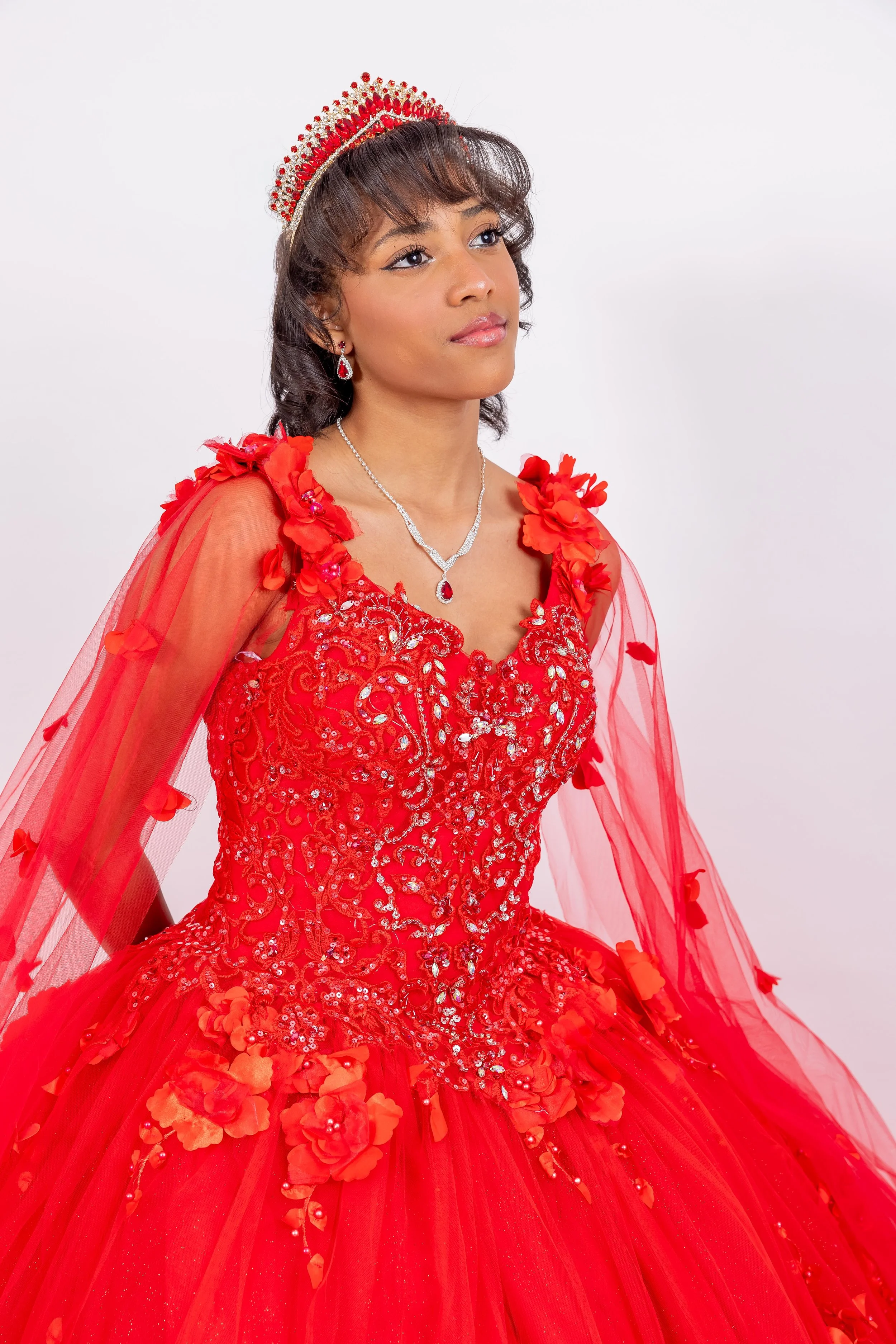 Joven mujer de cabello rizado, ataviada con vestido rojo de gala adornado con flores y lentejuelas, lleva una tiara y joyería, posando en un fondo blanco.
