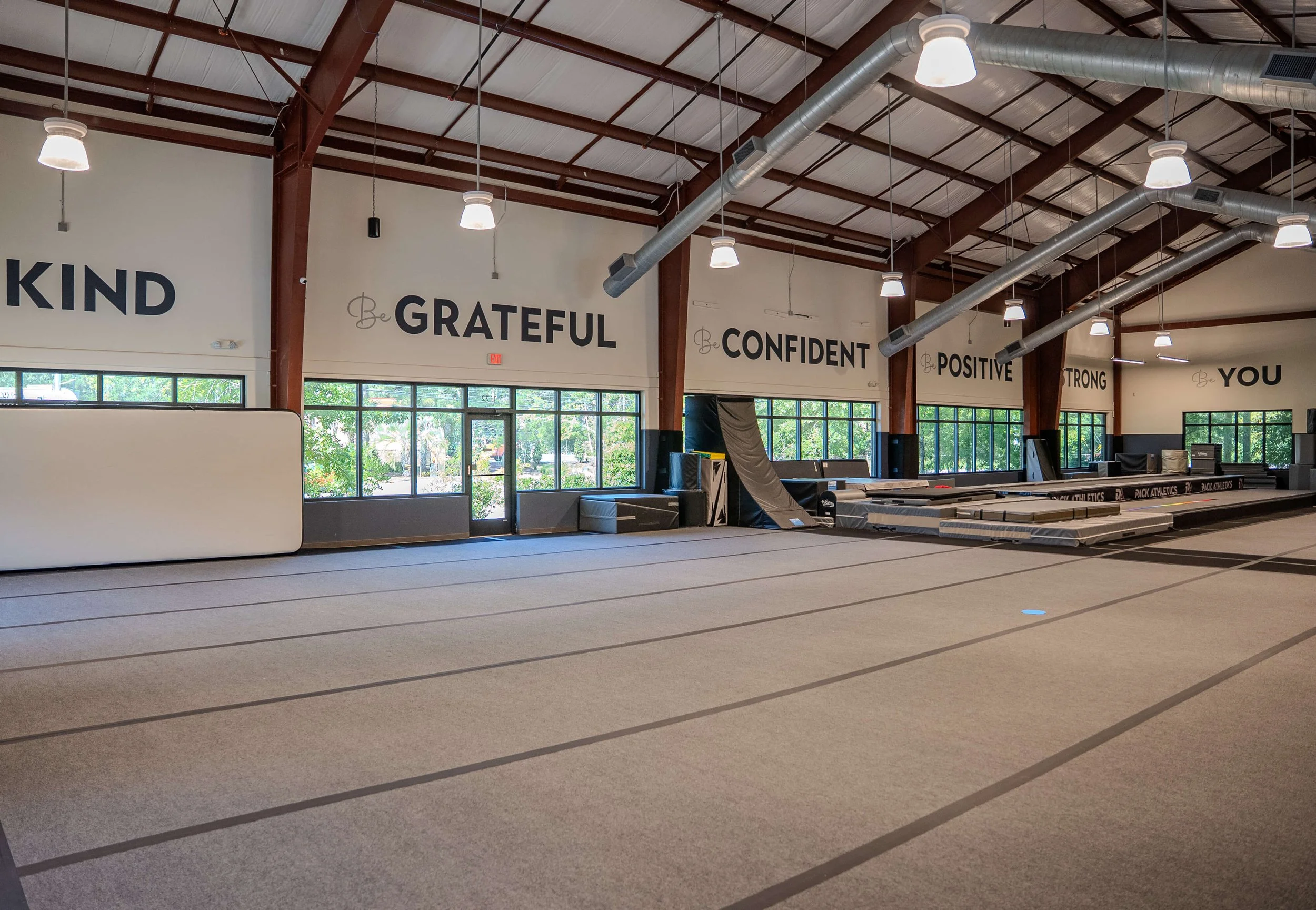 Empty gymnastics training facility with large open floor, mats, foam pits, and motivational words on the wall such as "Be GRATEFUL," "Be CONFIDENT," "Be POSITIVE," "Be STRONG," and "Be YOU." Large windows let in natural light.