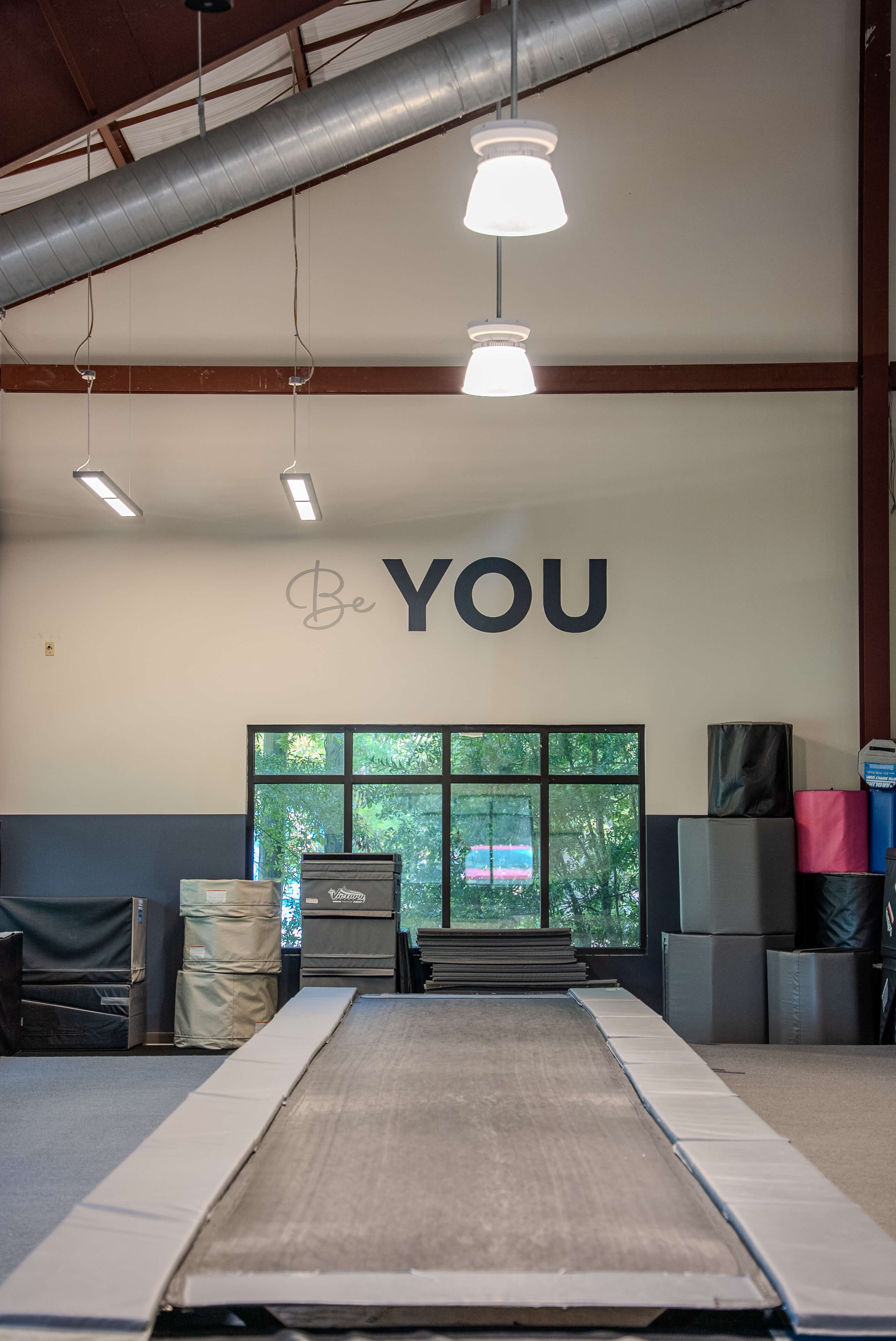 Indoor gym with a trampoline in the foreground and a wall with the words 'Be YOU' in large letters, windows showing trees outside, and various gym equipment stacked against the wall.
