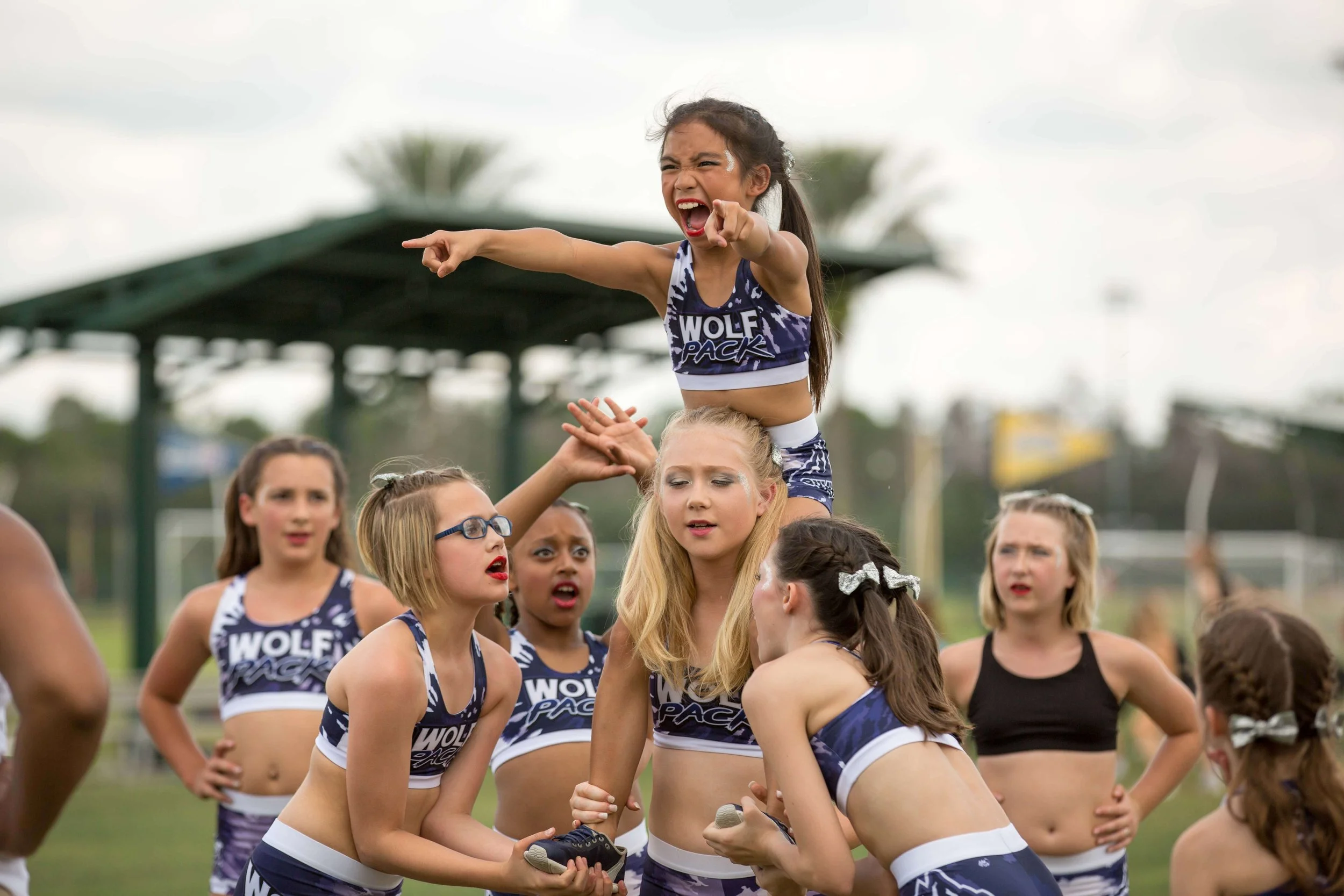 A group of young cheerleaders practicing outdoors, with one girl lifted by others, pointing and shouting, wearing matching cheer uniforms.