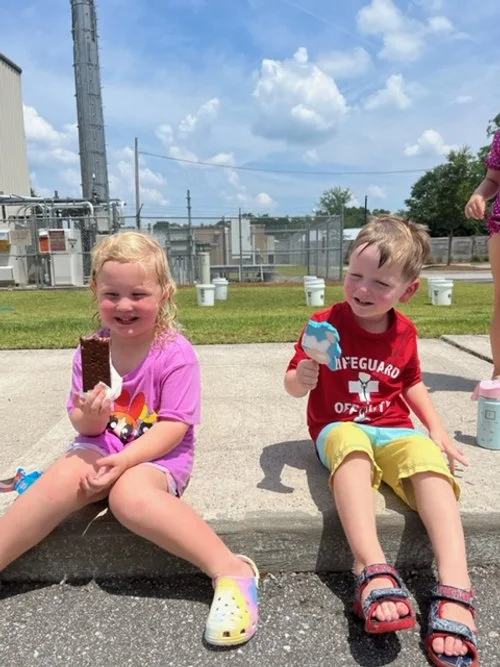 Two young children sitting on a concrete curb eating ice cream, with a grassy area, electrical infrastructure, and a blue sky with clouds in the background.