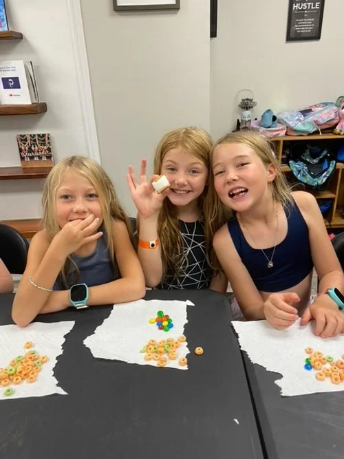 Three young girls sitting at a table with cereal and fruit loops, smiling, with one girl holding a piece of cereal.