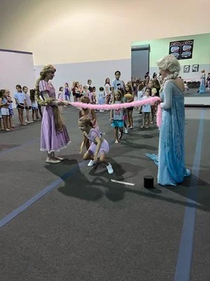 Two women dressed as Disney princesses, Anna and Elsa, entertain young children at a party with a pink cotton candy machine. Children and adults are gathered in a large room with play and food stations.