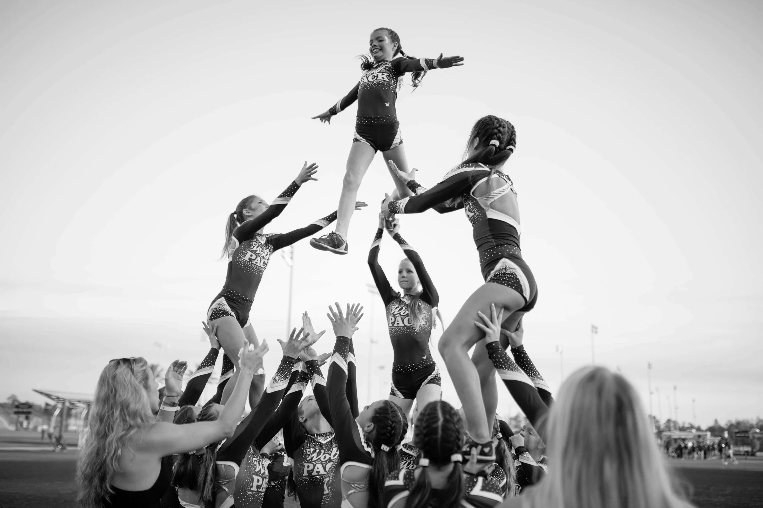 Cheerleaders in uniform performing a cheerleading stunt with one girl elevated above the others, against an outdoor sports field background.