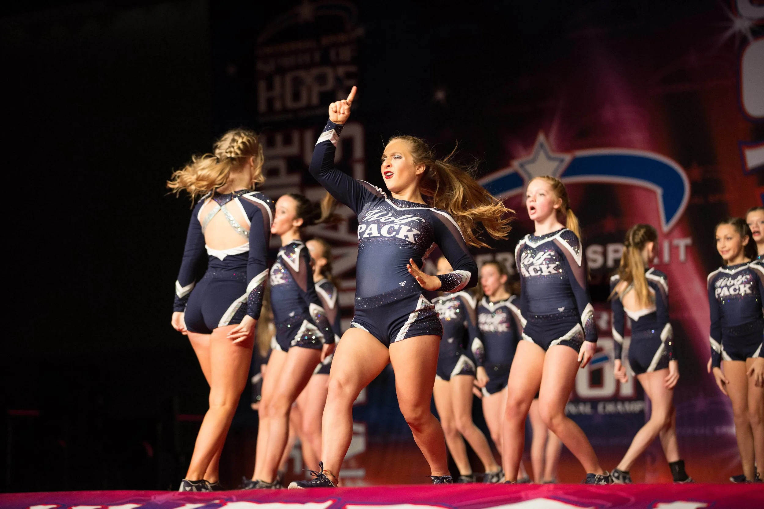 A group of cheerleaders performing on stage in matching navy blue and white costumes, with some raising their arms and others dancing.