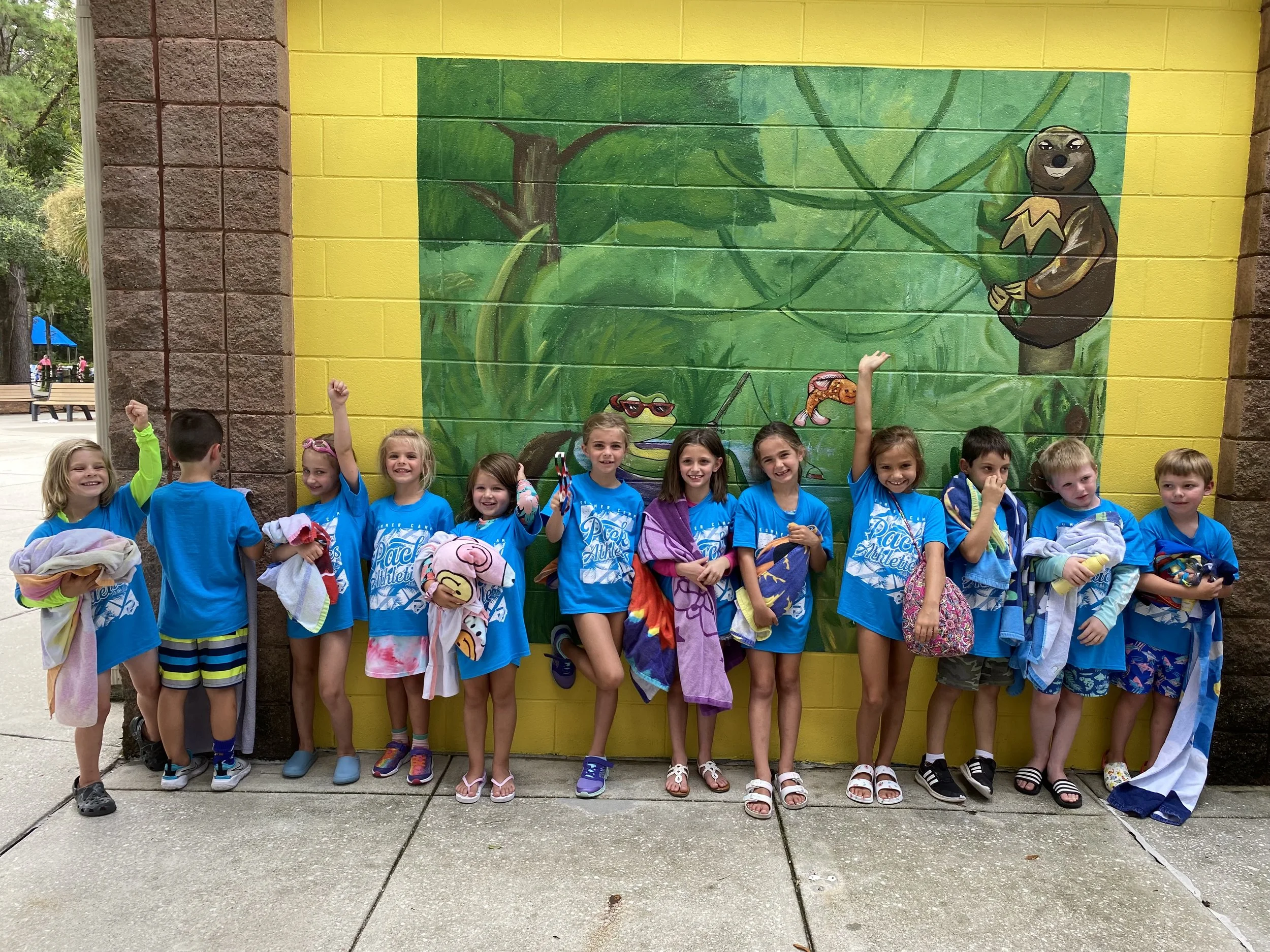 Group of children in blue shirts standing in front of a colorful jungle mural, smiling and holding towels and swim gear outside on a concrete sidewalk.