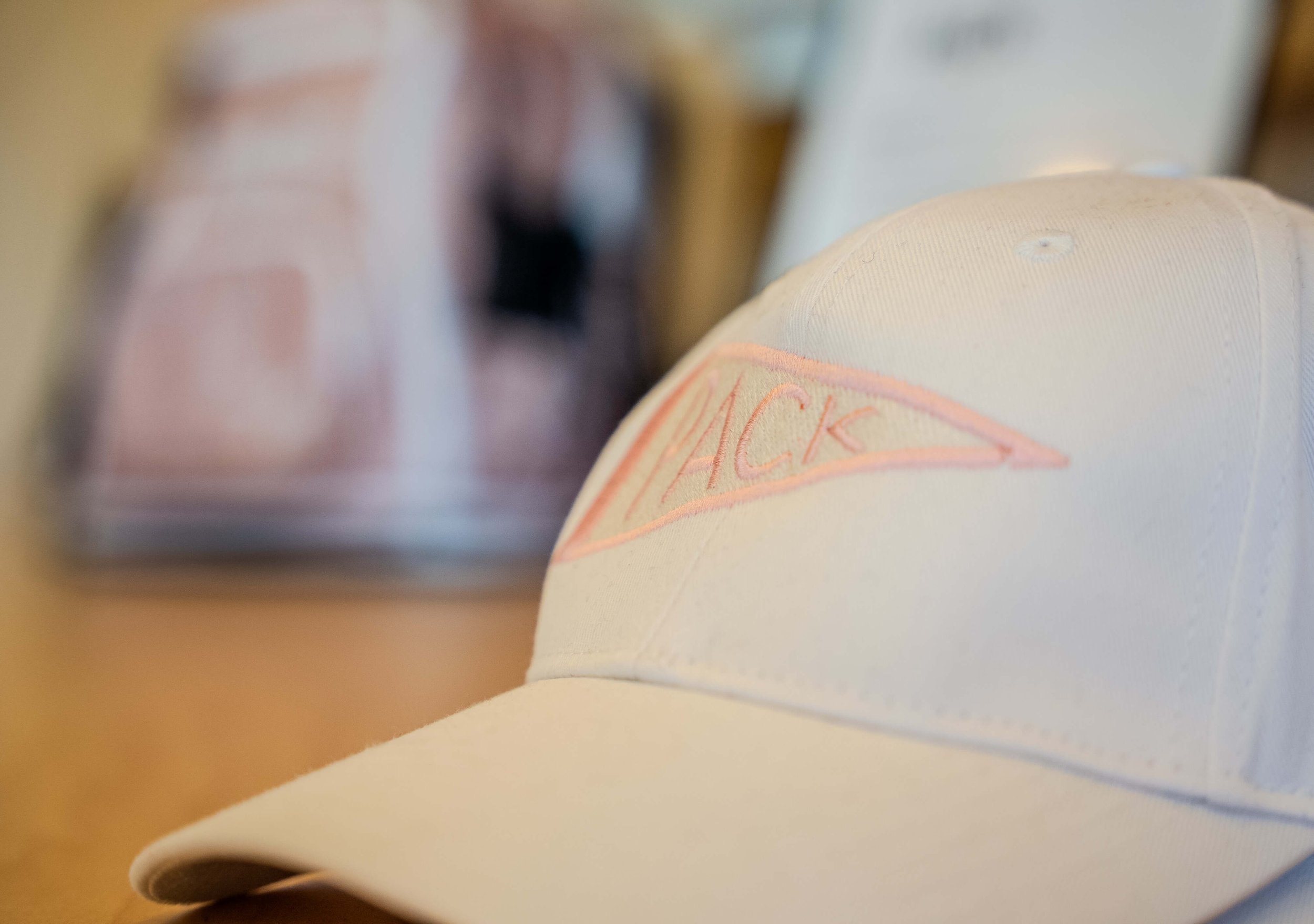 A light-colored baseball cap with the word "HACK" embroidered in pink on the front. The cap is placed on a surface, with blurred background objects.
