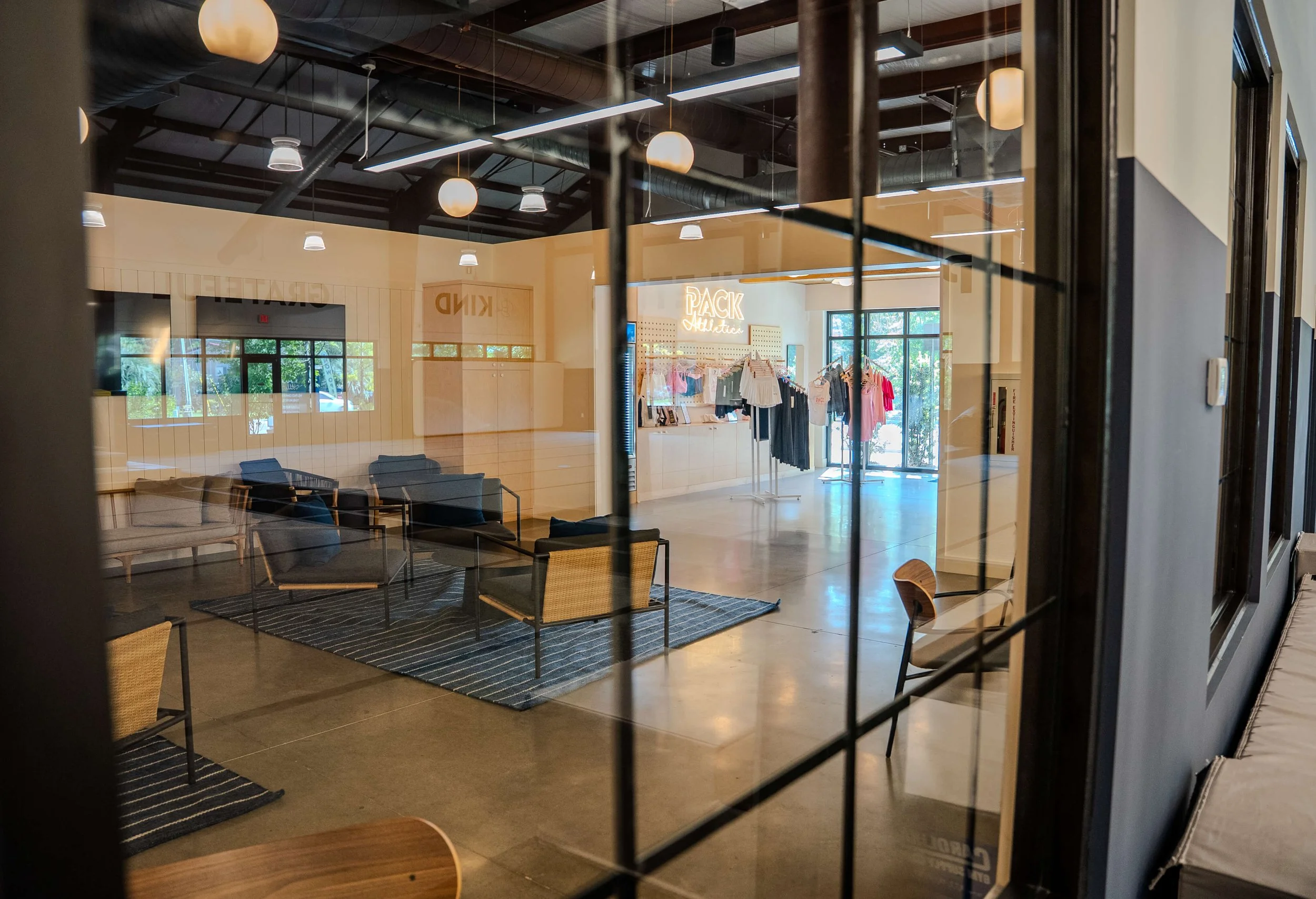 Interior view of a clothing retail store seen through a glass window with grid pattern, showing seating area, a clothing display with a neon sign, and large windows.
