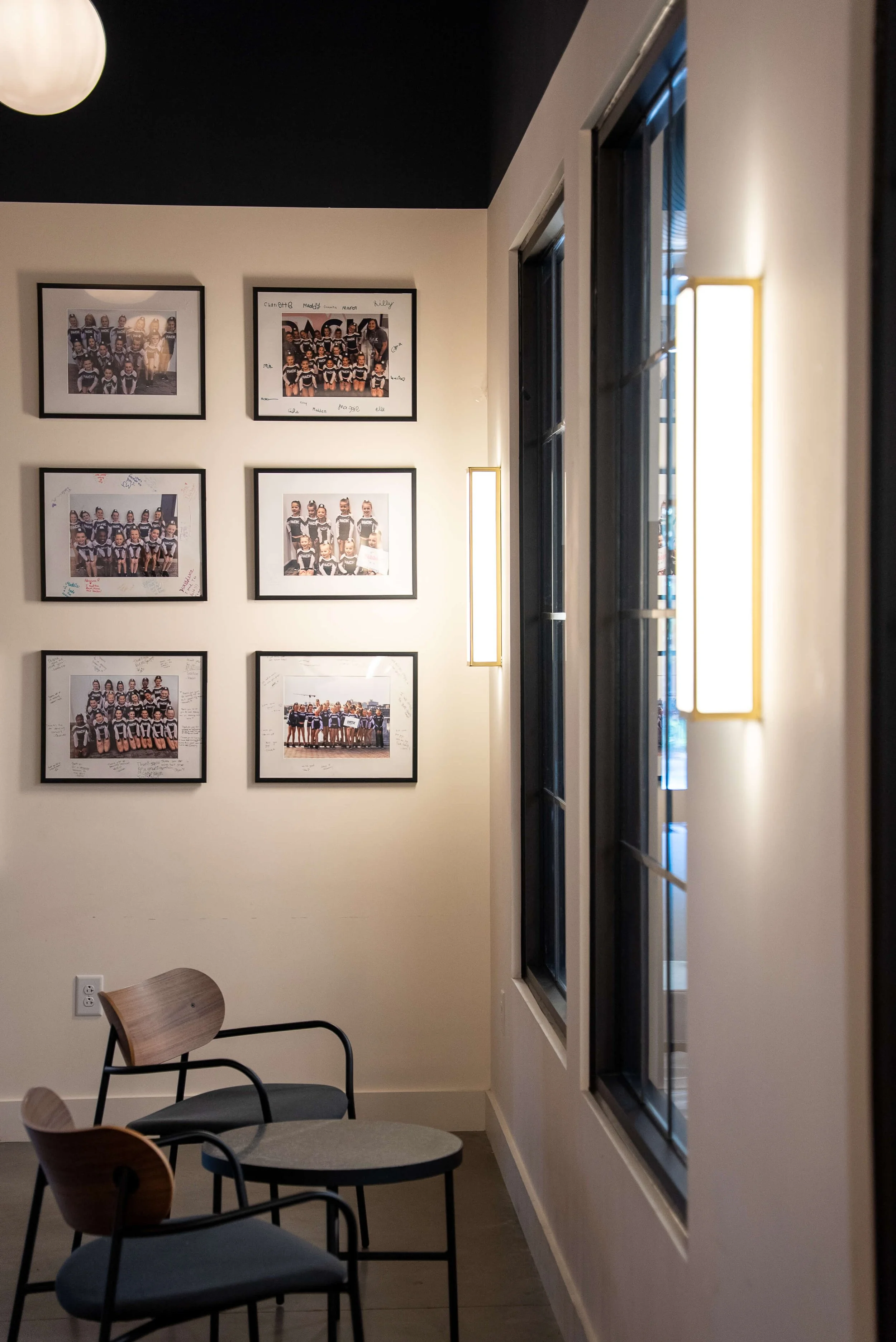 A corner of a room with six framed group photos of kids on the wall, two modern chairs, a small round table, beige walls, large windows, and wall-mounted light fixtures.