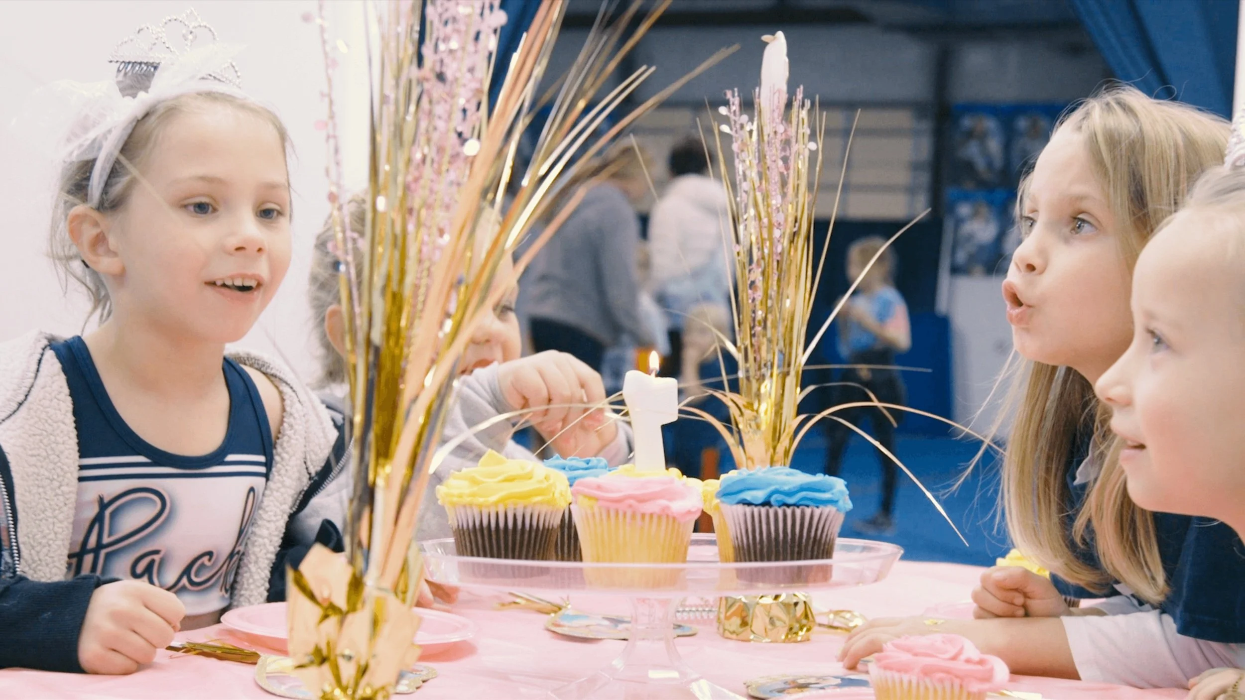Four young girls celebrating a birthday, gathered around a table with colorful frosted cupcakes and a lit candle, decorated with gold and pink party decorations