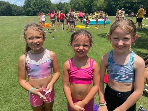 Three young girls standing on a grassy field during a pool party, with people, pool, and trees in the background.