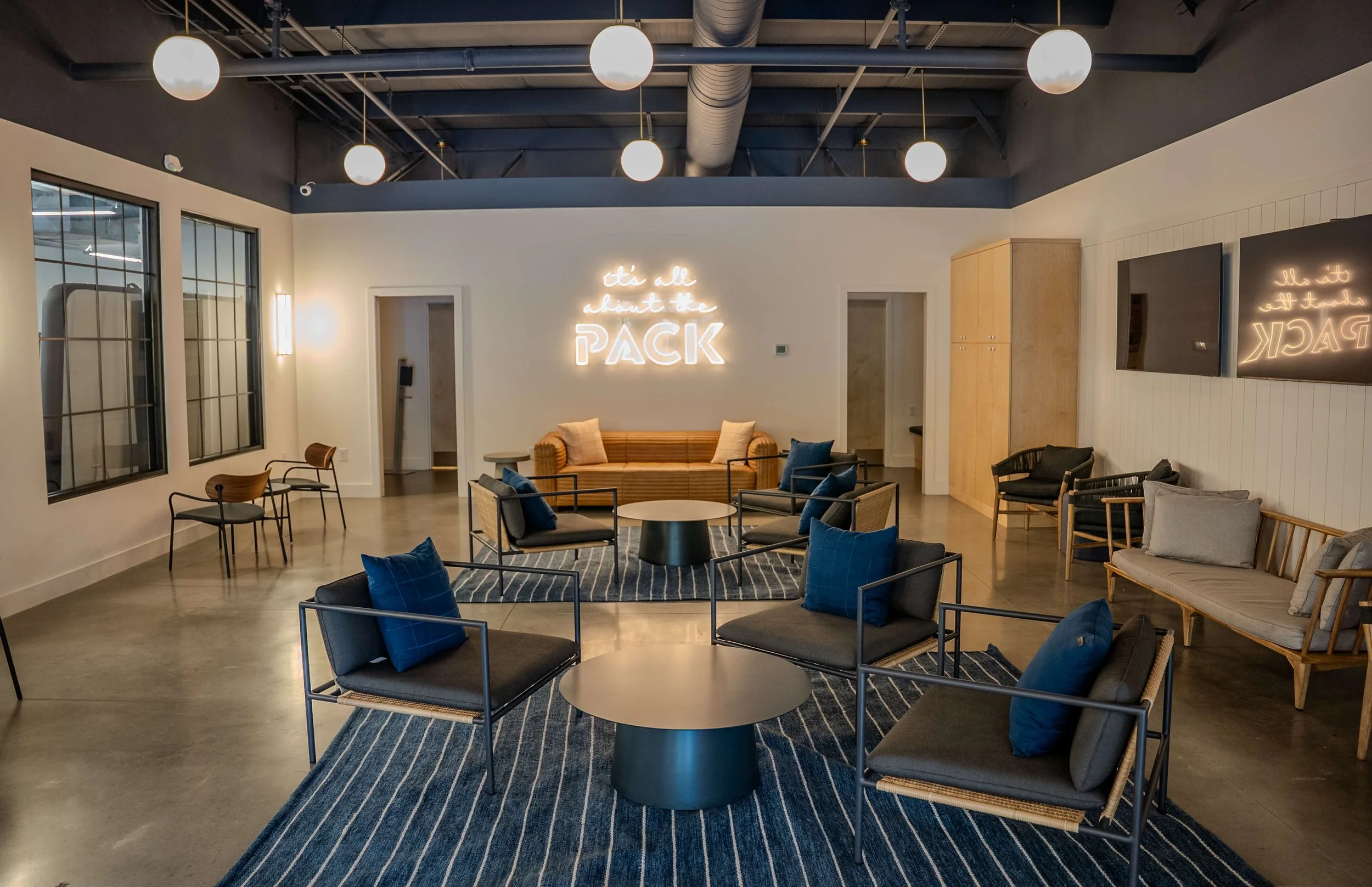 Modern lounge area with black and blue chairs, beige sofas, circular coffee tables, striped rugs, and a neon sign on the wall that says 'it's all about the PACK.'