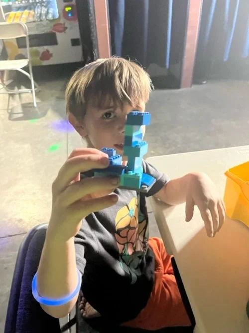 Child sitting at a table, holding up a LEGO creation, with a snack container on the table, in a room with vending machines and chairs in the background.