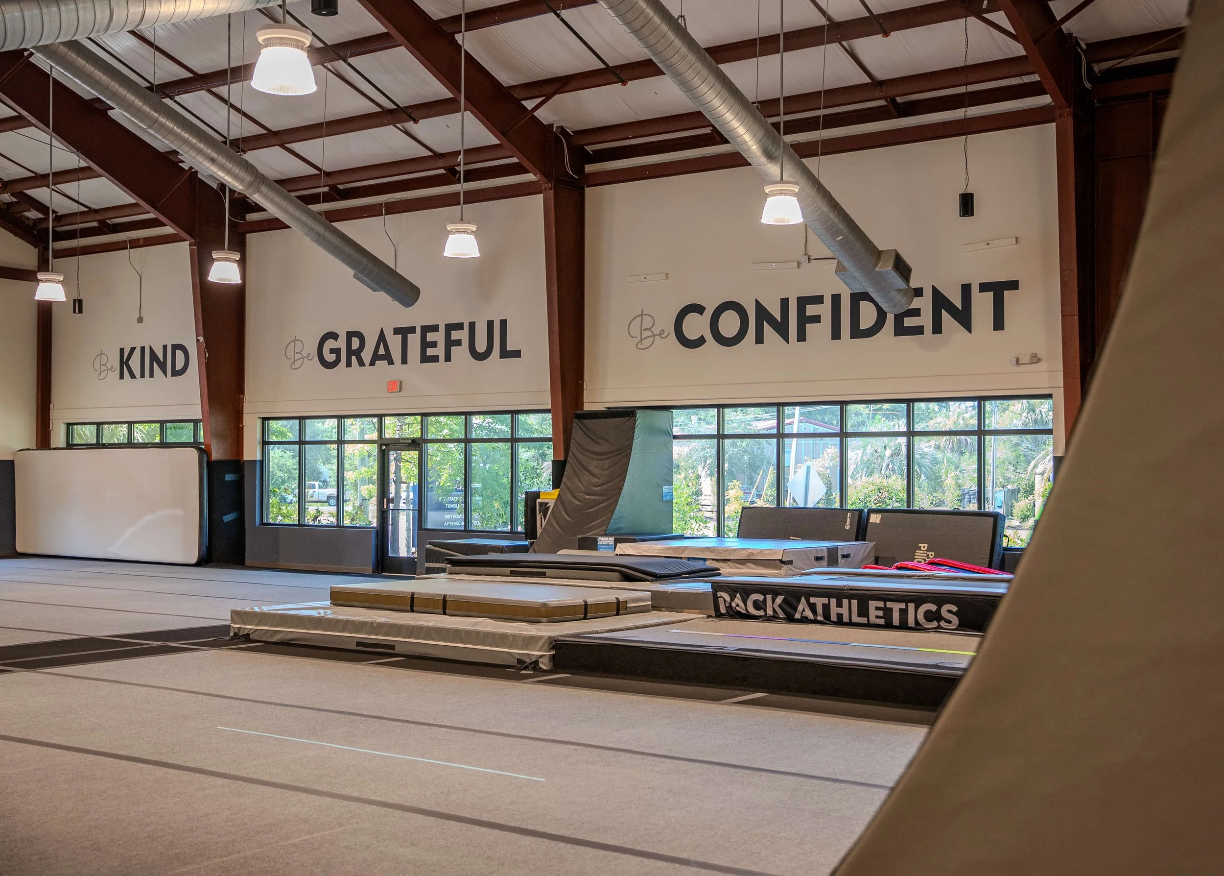 Inside a gym with large motivational signs on the wall: 'Be KIND', 'Be GRATEFUL', 'Be CONFIDENT'. There are gym mats and equipment, with windows showing trees outside.