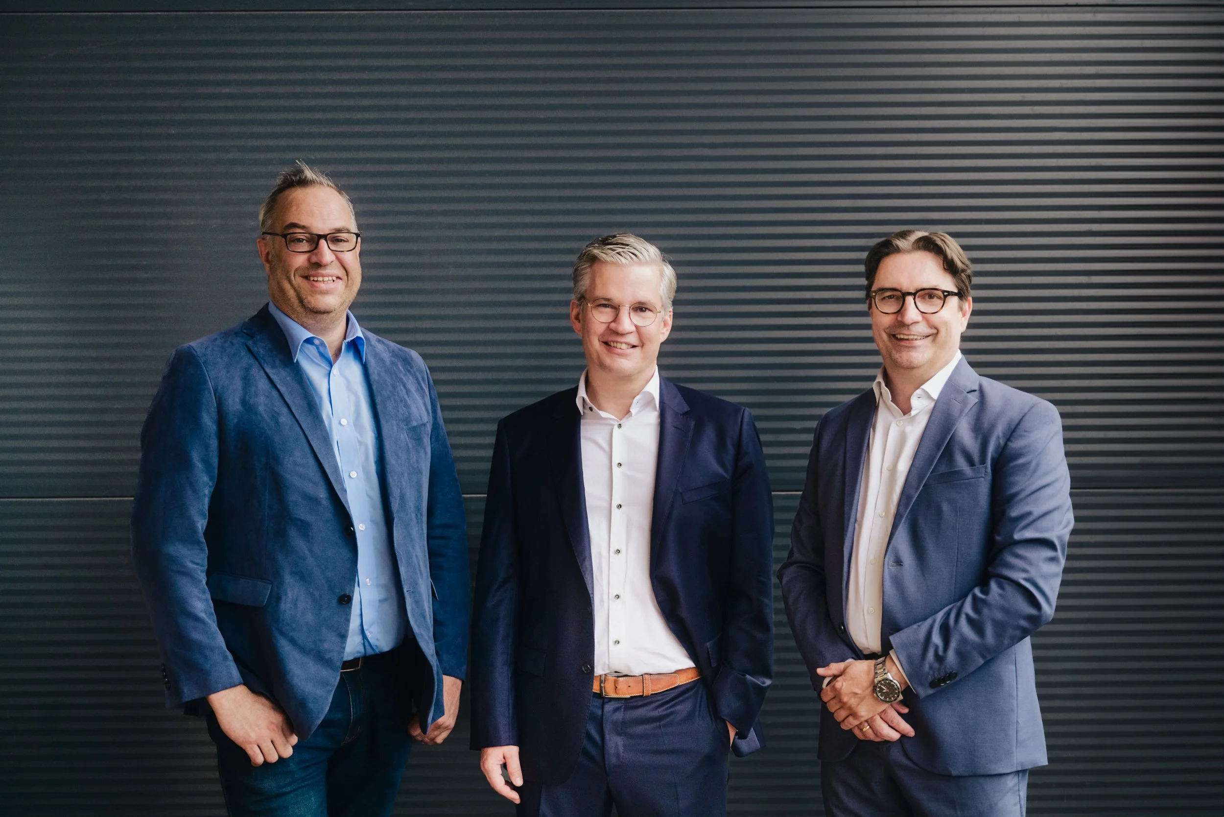 Drei Männer in formeller Kleidung stehen vor einer schwarzen Wand und lächeln in die Kamera.