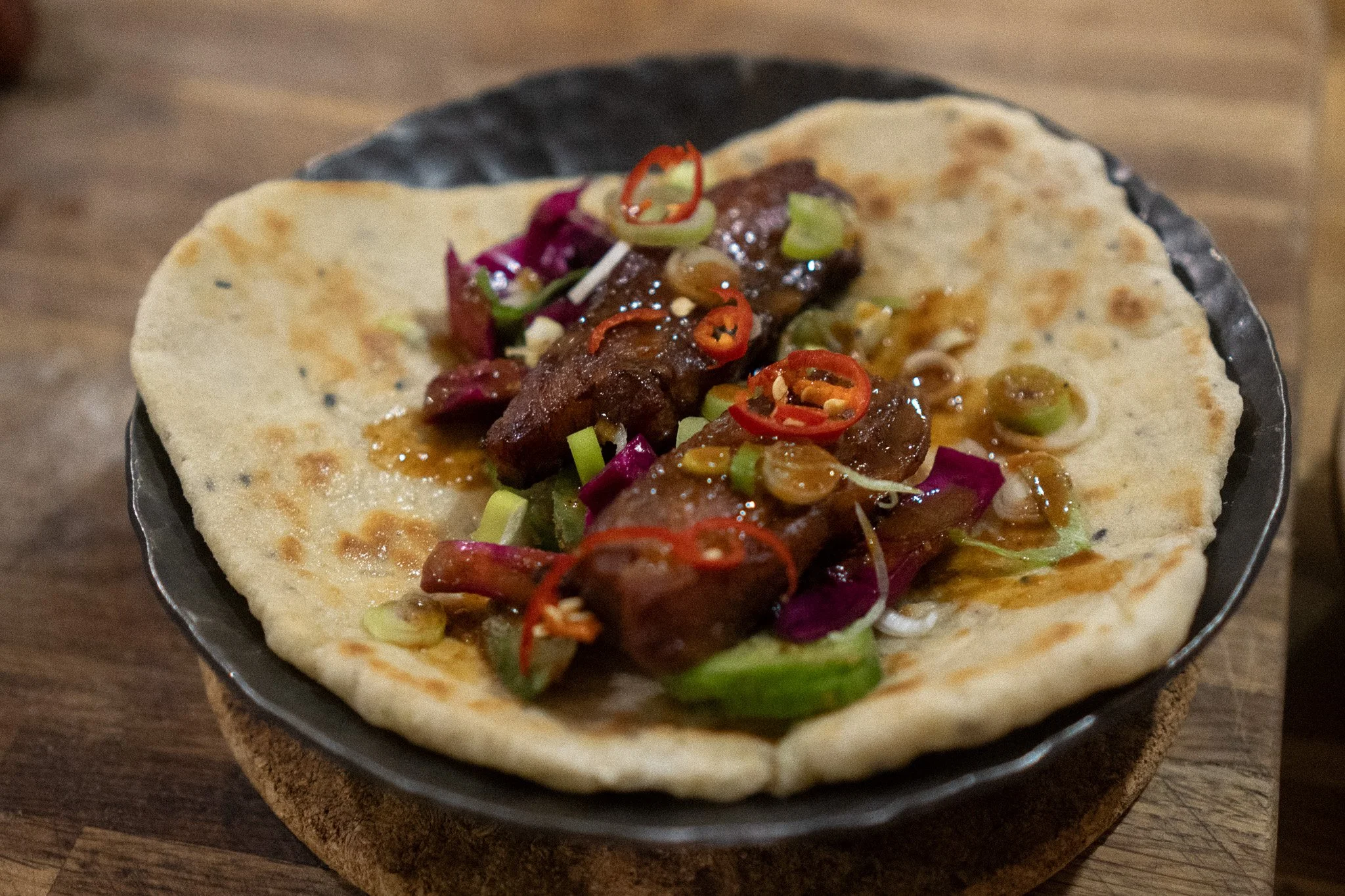 Close-up of a taco with beef, chopped green onions, sliced red chili peppers, and purple cabbage in a black bowl on a wooden surface.