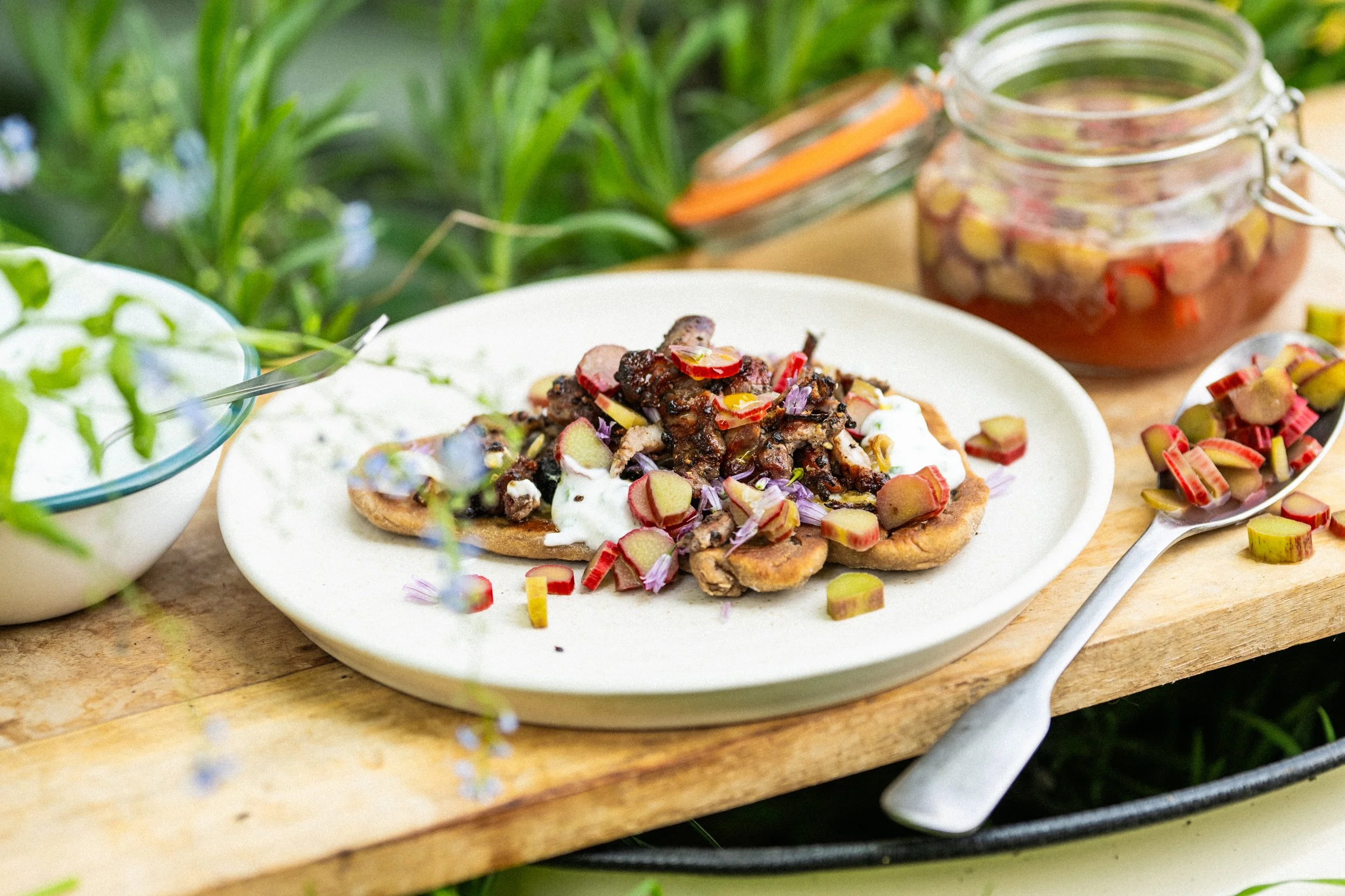 Open-faced sandwich with dark meat, chopped herbs, and sliced rhubarb on a white plate, with a jar of rhubarb and a spoonful of sliced rhubarb nearby, set on a wooden surface outdoors with green foliage in the background.