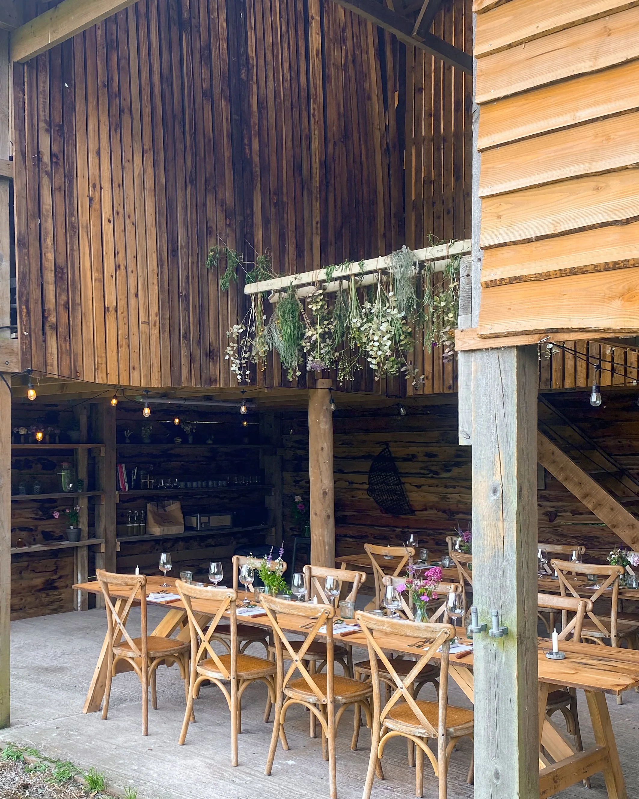 Outdoor dining area with wooden tables and chairs set for a meal, decorated with small flower arrangements, under a wooden covered patio with string lights and a wall decorated with hanging greenery.