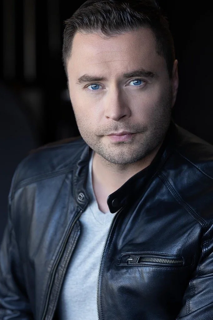 Close-up portrait of a man with blue eyes, short dark hair, wearing a black leather jacket and a light gray shirt, against a dark background.
