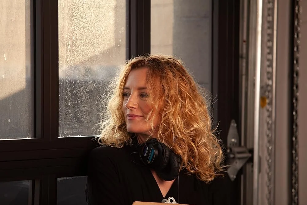 A woman with curly blonde hair and black headphones around her neck, looking thoughtfully out a window with rain droplets, in an industrial-style setting.