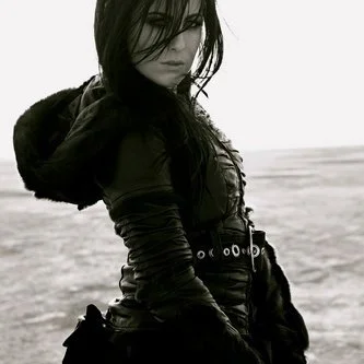 A young woman with long black hair wearing a black jacket and belt standing outdoors on a beach with a cloudy sky.