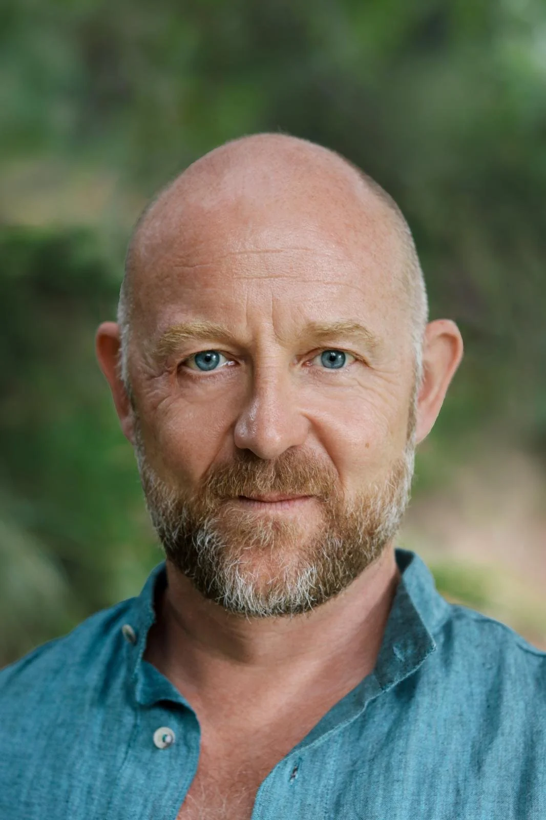 Close-up of a middle-aged man with a bald head, blue eyes, and a beard, wearing a blue button-up shirt, outdoors with a blurred green background.