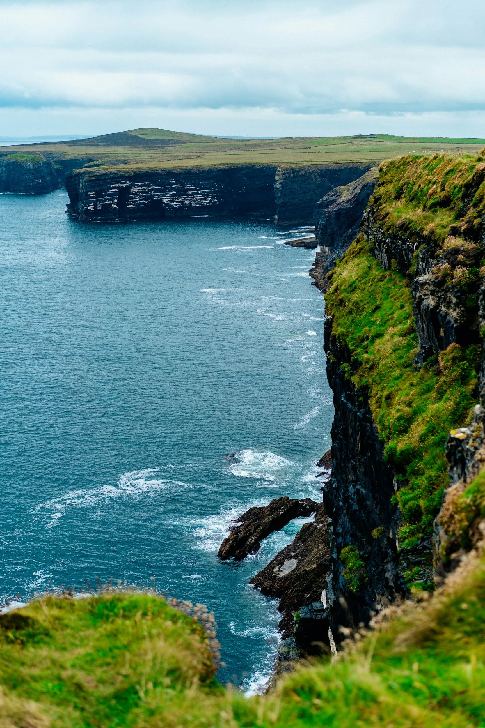 Cliffs overlooking the ocean, with grassy tops and rocky faces, under a cloudy sky.