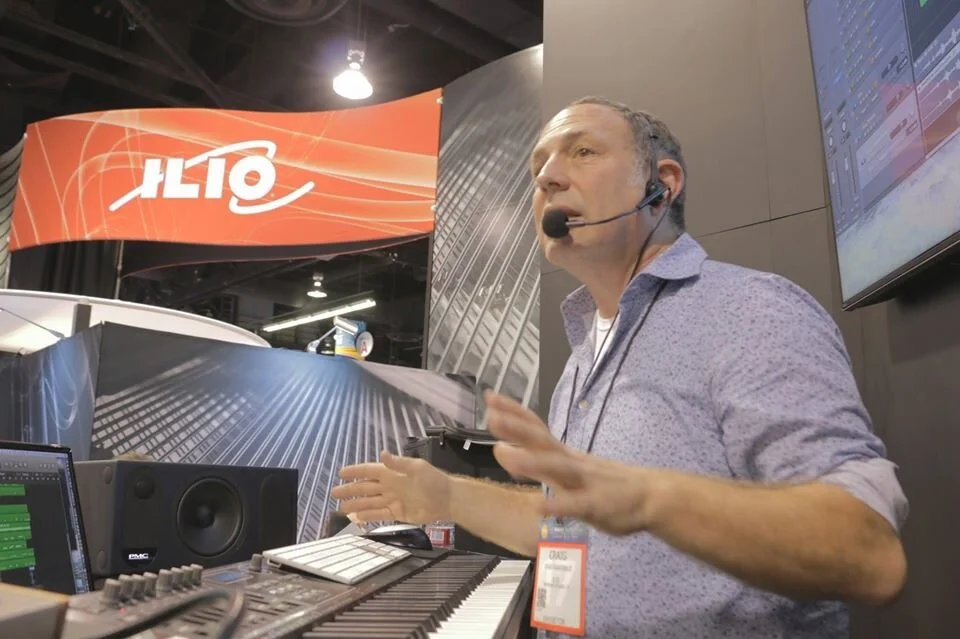 A man wearing a headset microphone gestures with his hands as he speaks at a booth with music production equipment, including a keyboard, monitor speaker, and a computer. A large screen displays audio editing software nearby, and a red and white banner with the word 'ALT' is visible above.
