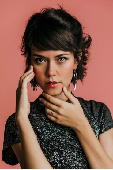 A woman with dark wavy hair and piercing blue eyes posing against a pink background.