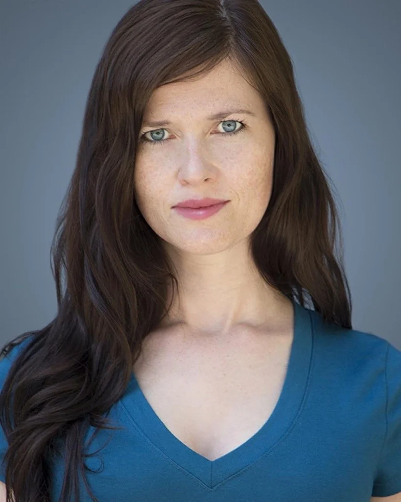 A woman with long wavy brown hair, blue eyes, and freckles, wearing a blue V-neck shirt, posing against a plain grey background.