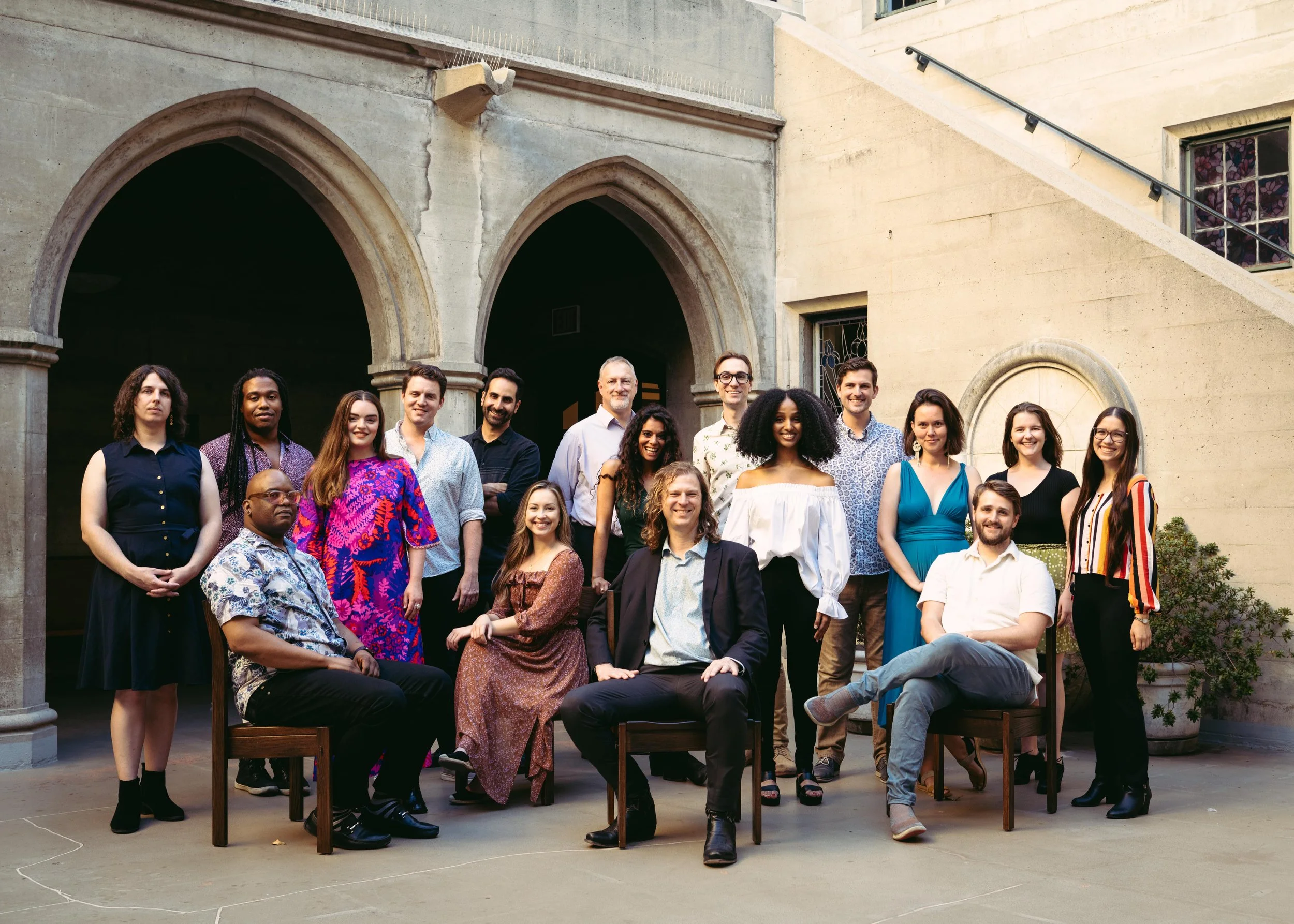Group of 16 diverse people posing outside a building with arches and stained glass windows, with some seated and others standing, smiling at the camera.