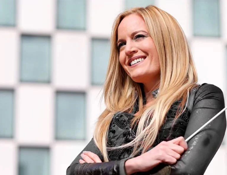 A blonde woman smiling with arms crossed outside in front of a modern glass building.
