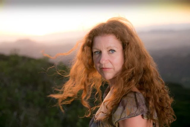 A woman with long, curly red hair outdoors at sunset or sunrise, looking directly at the camera with a serious expression.