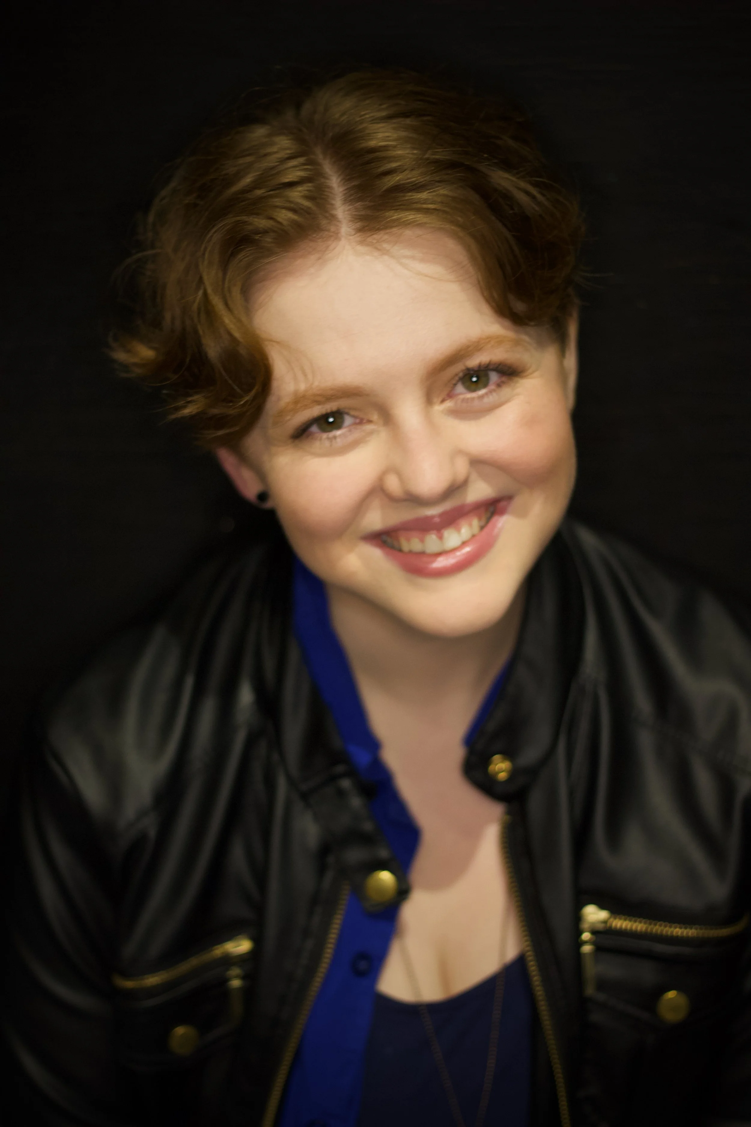 A woman smiling with short, wavy hair wearing a black leather jacket and a blue shirt against a dark background.